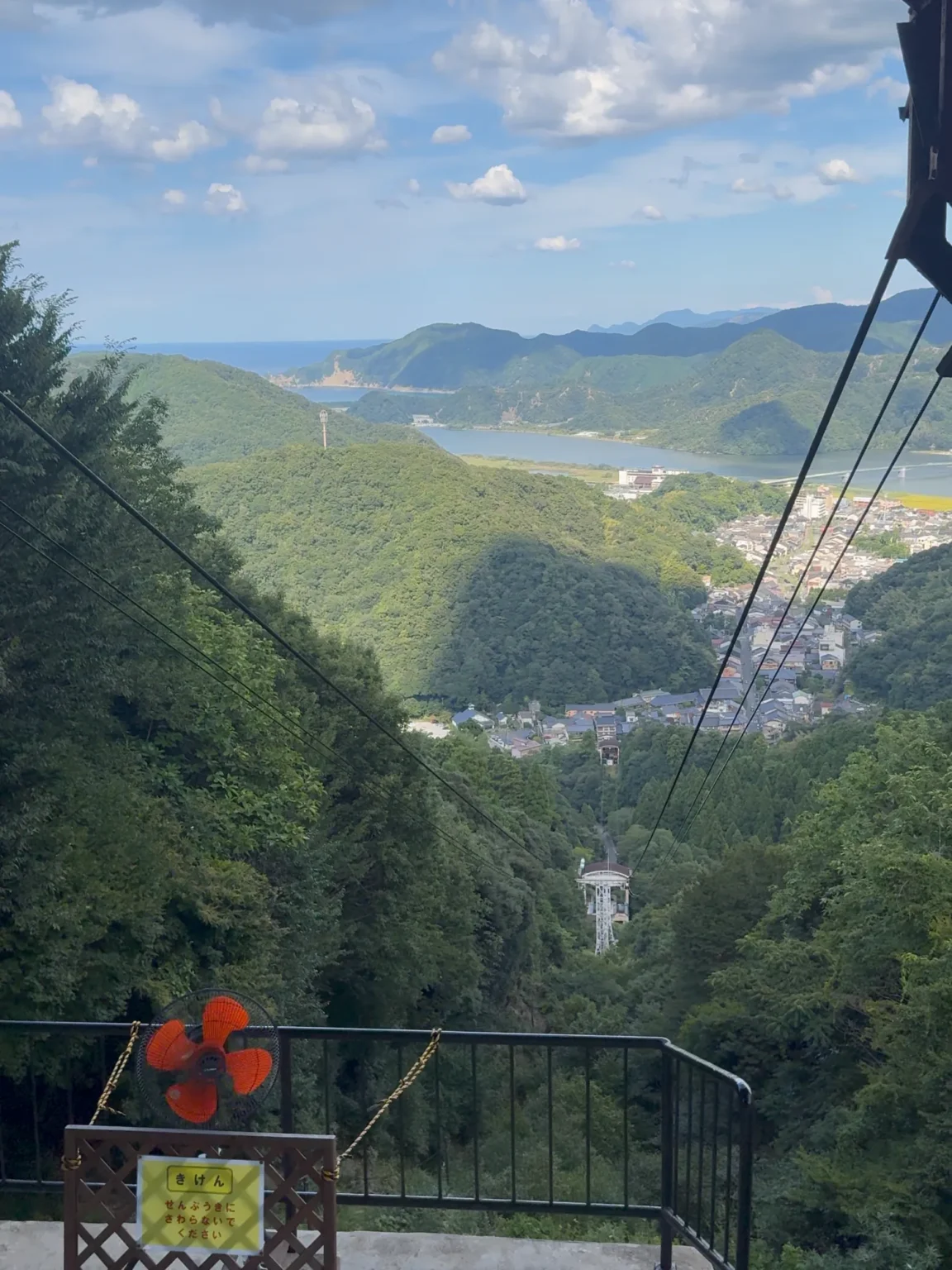 Looking down the ropeway cables from the top station on Daishi-yama: a lattice tower in the forest, the rooftops of Kinosaki Onsen in the valley, the Maruyama River and green hills, with an orange fan and a Japanese warning sign at the platform railing