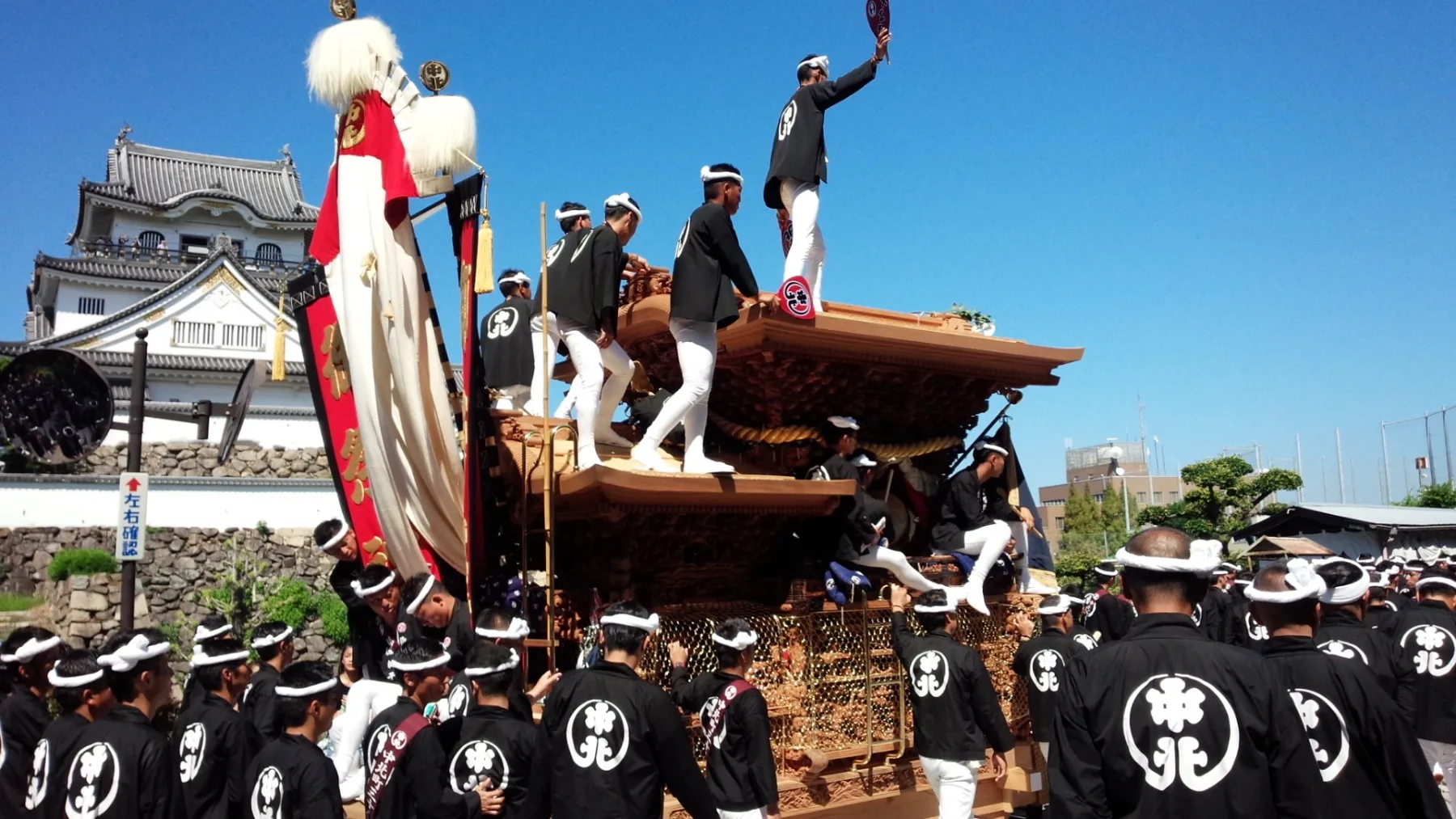 A danjiri float with its roof rider standing on top, passing below the keep of Kishiwada Castle