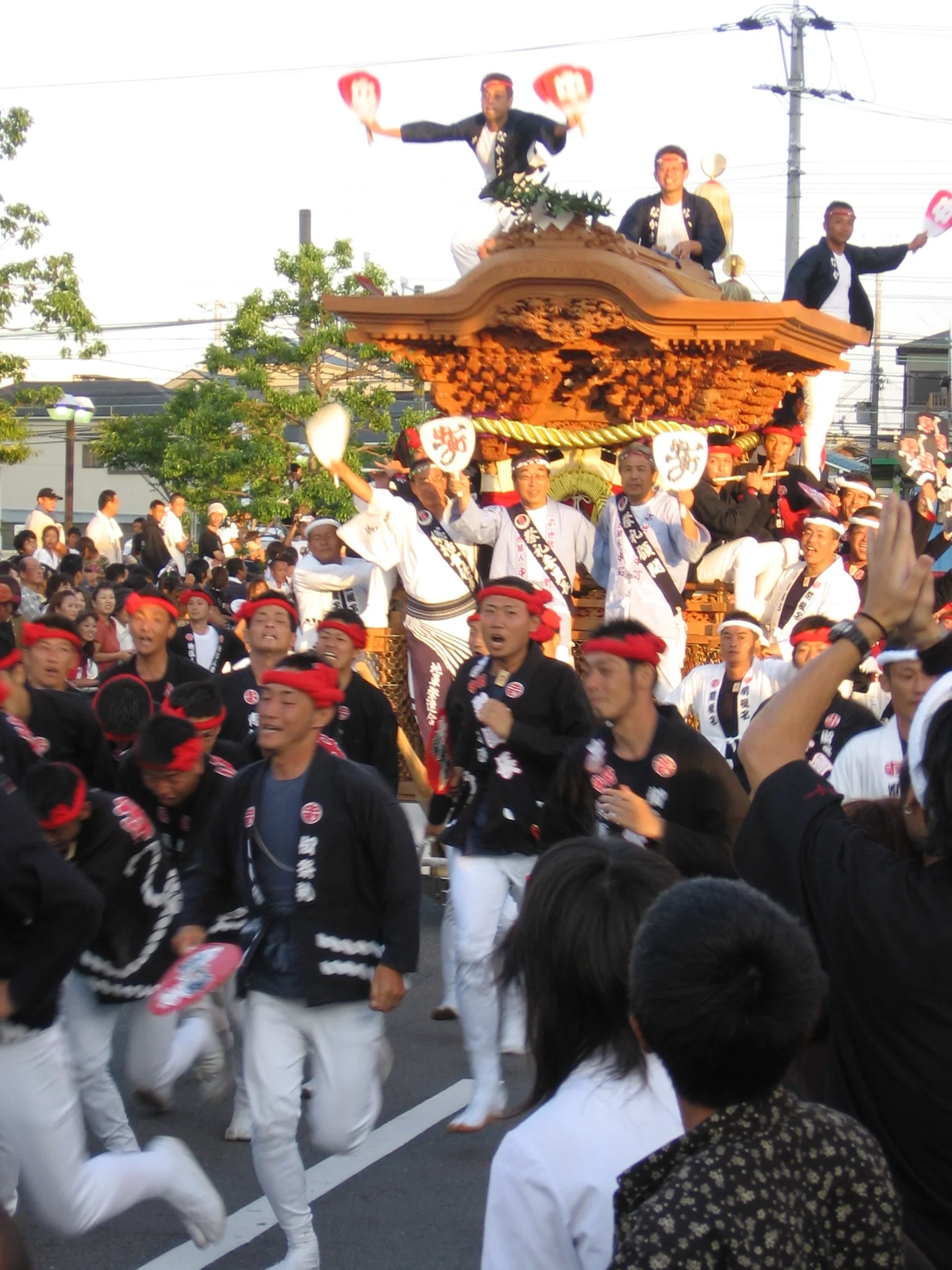 The daiku-gata roof dancer holding fans above a danjiri float as the pulling crew runs it through a Kishiwada street