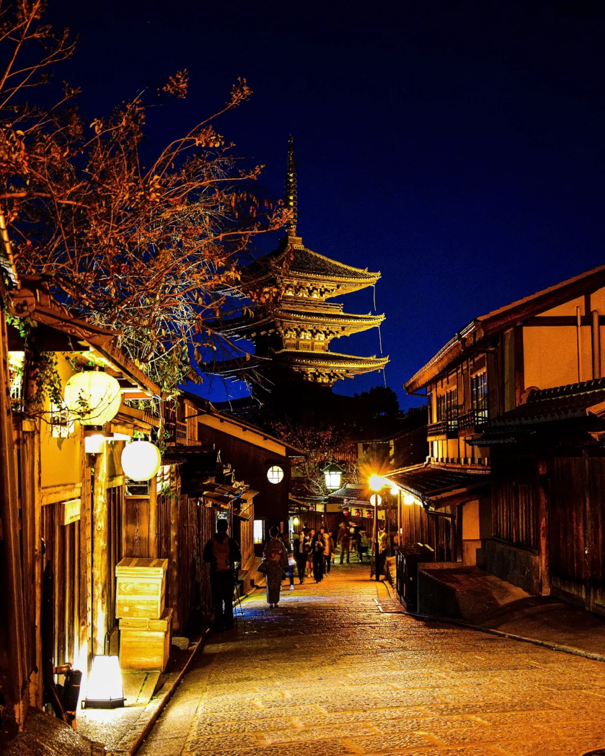 The illuminated Yasaka Pagoda rising above a stone-paved lane of wooden townhouses in Kyoto at night, round lanterns glowing beside the doors and a few people walking up the lane