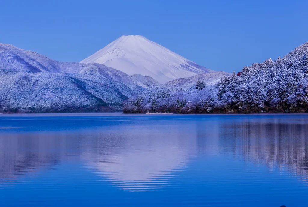 Snow-dusted Mount Fuji rising beyond frost-covered trees at Lake Ashi in Hakone, its white cone mirrored in the still blue water of the lake on a clear winter morning