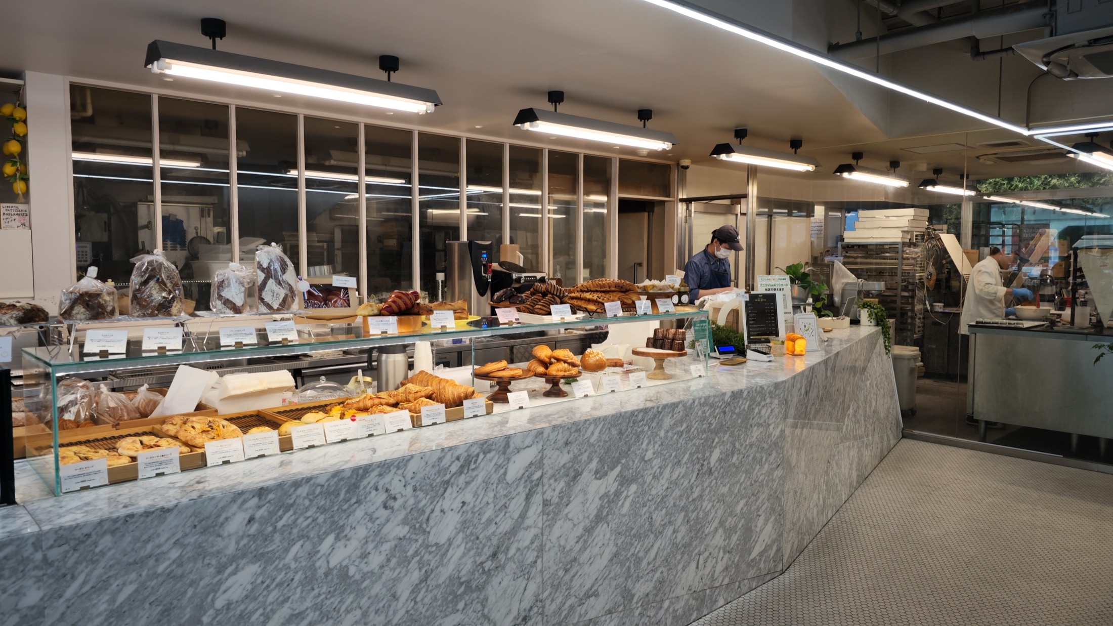 The ground-floor bakery counter at Liberte Kichijoji, a long marble counter with glass cases of viennoiserie and loaves, staff working behind it and bread racks in the open workshop beyond