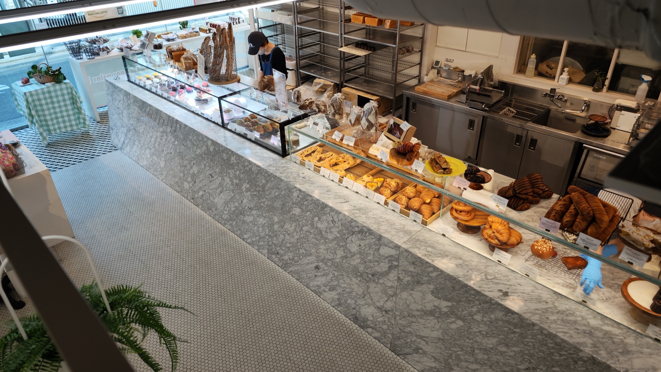 The Liberte Kichijoji bakery counter seen from the second-floor cafe above, showing the marble counter, pastry and cake cases, white hexagonal tile floor and a staff member serving at the till