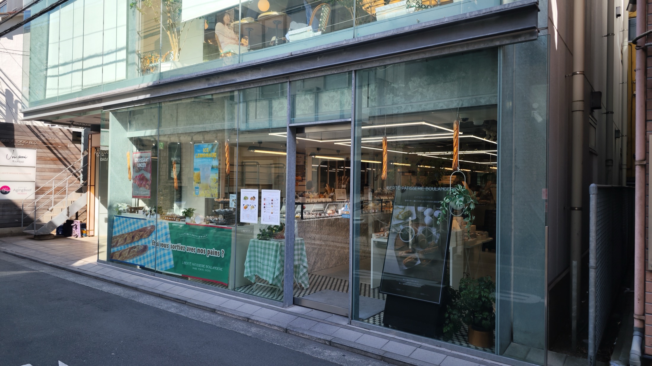 The fully glazed two-storey shopfront of Liberte Patisserie Boulangerie in Kichijoji on a narrow side street, with baguettes hanging in the window, a green gingham banner and the upstairs cafe visible through the glass above