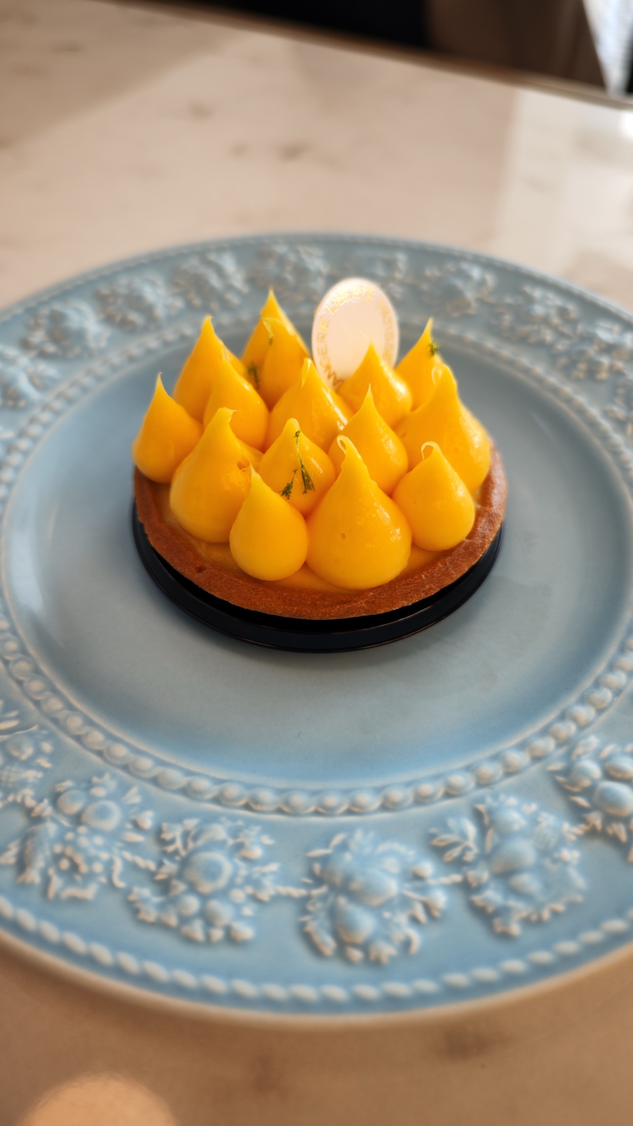 A tarte citron at Liberte Kichijoji, covered in piped yellow lemon cream teardrops with fine green lime zest and a white oval brand plaque on top, served on its black base on a pale blue relief plate on a marble table