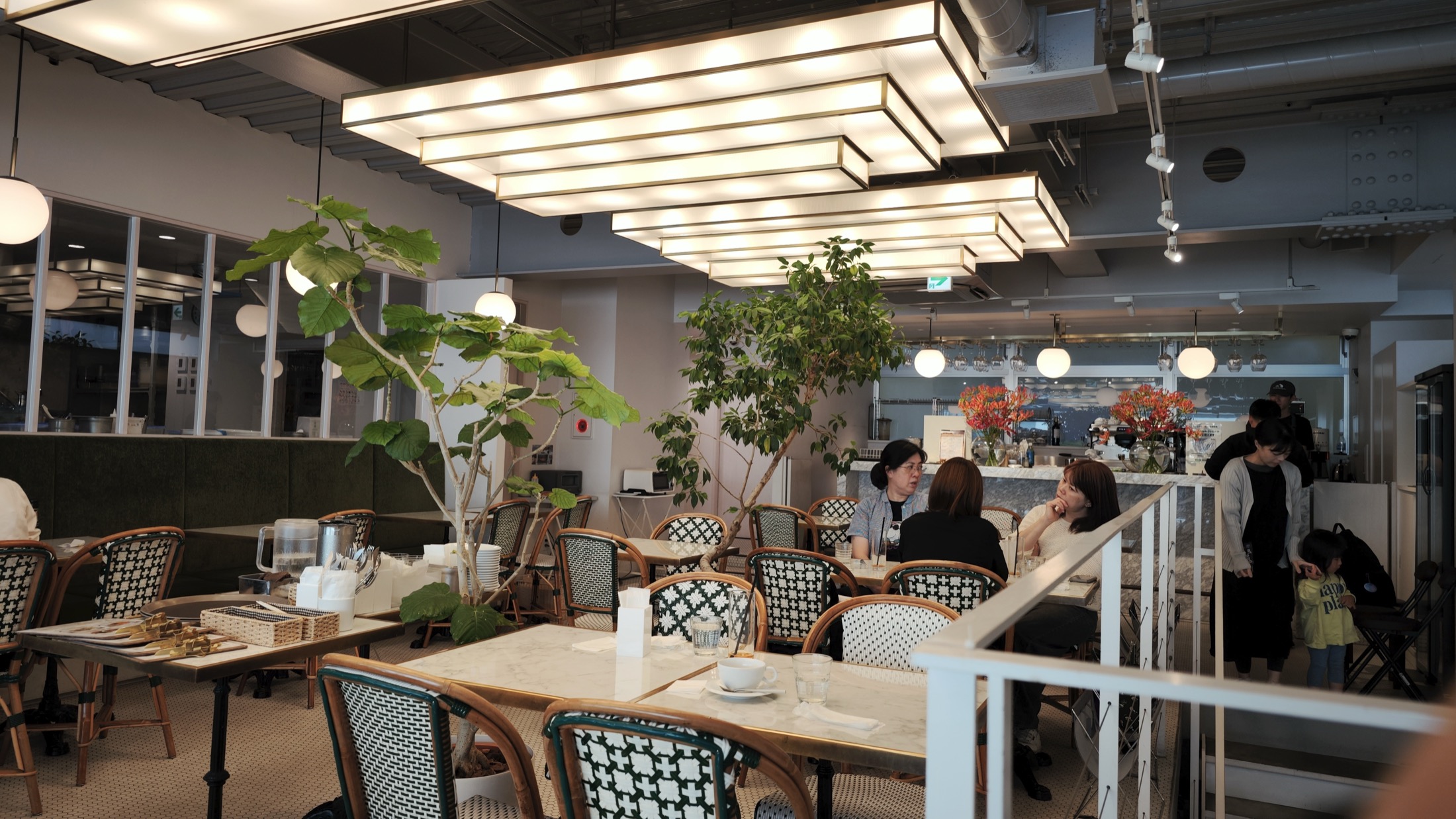 The second-floor cafe at Liberte Kichijoji, with rattan bistro chairs woven in green and white, marble-topped tables, indoor trees reaching toward stepped rectangular ceiling lights, and customers seated along the window