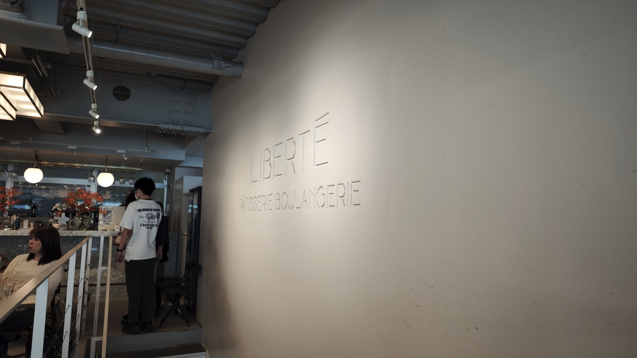 The words LIBERTE PATISSERIE BOULANGERIE cut into a plain plaster wall on the second floor at Kichijoji, with the cafe seating and pendant lights visible to the left