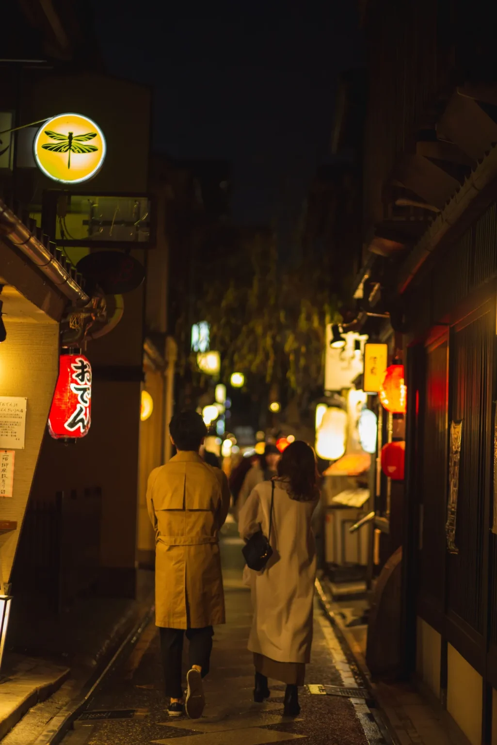 Two people seen from behind walking down a narrow lane at night, one in a belted beige trench coat and one in a long pale coat, with red and white lanterns and lit shop signs on either side