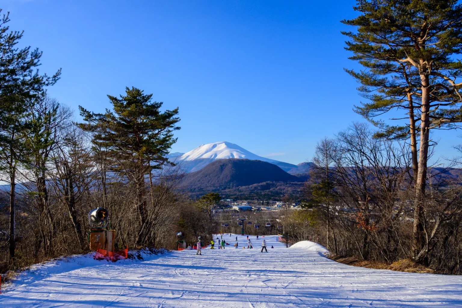 A groomed ski piste in Nagano dropping away toward the snow-capped cone of Mount Asama under a deep blue winter sky, with pine trees framing both sides of the run and skiers small in the distance