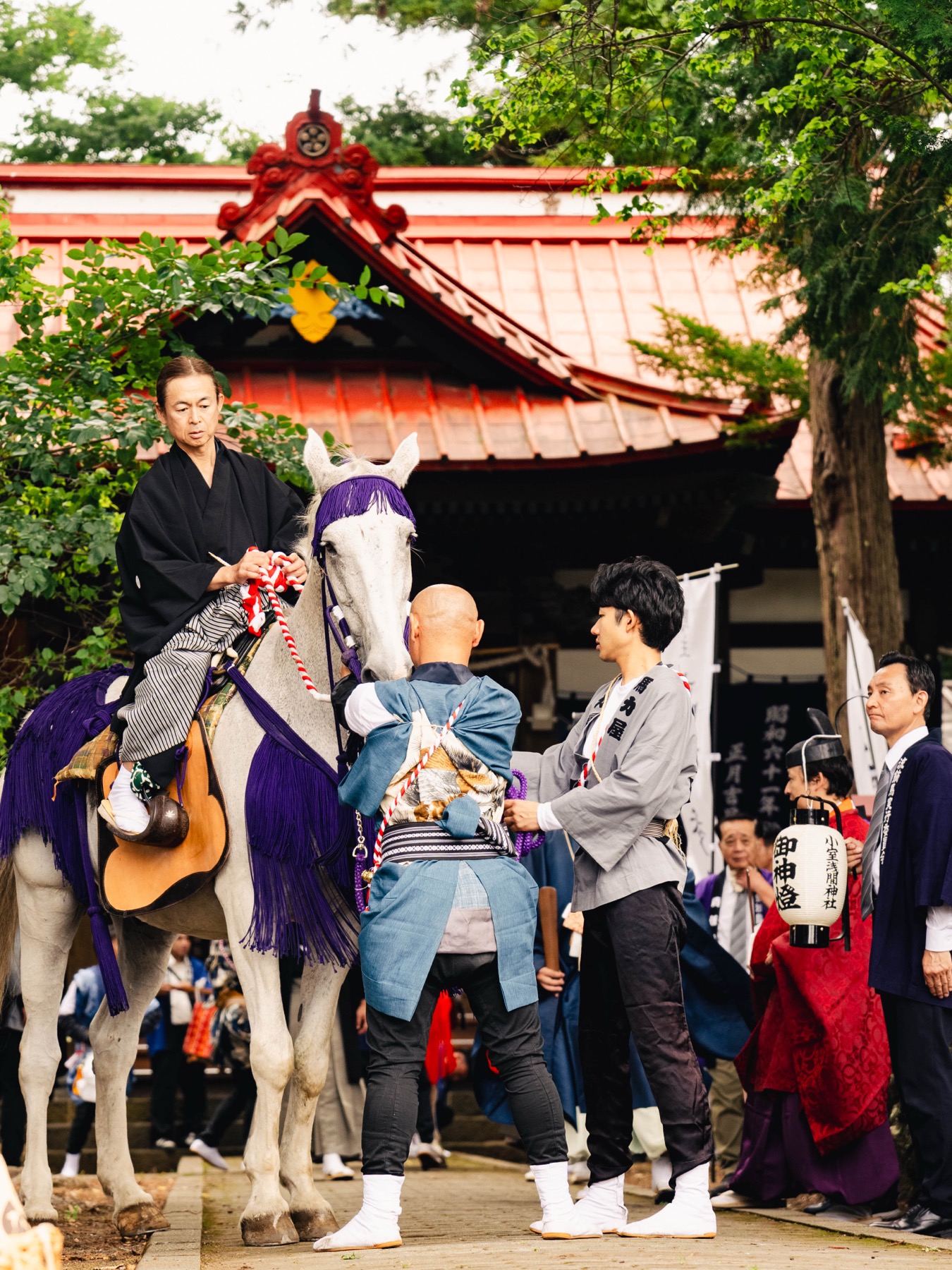 A rider in black formal dress sits on a white horse in front of the vermilion worship hall of Omuro Sengen Shrine while attendants in blue happi steady the horse and adjust the tack