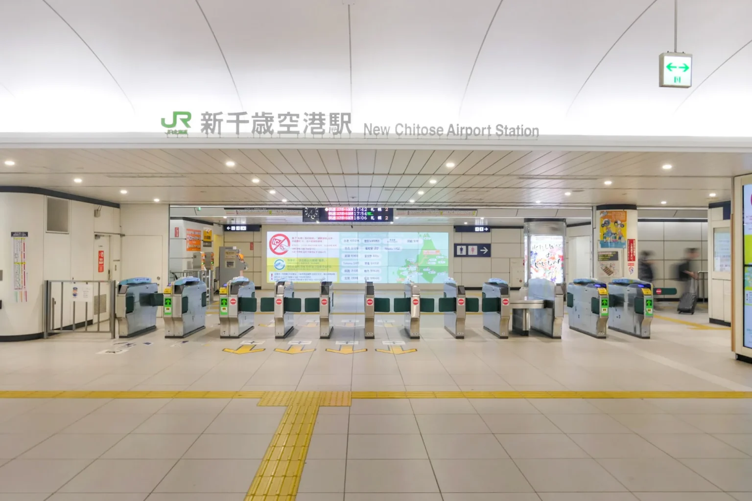 The JR ticket gates of New Chitose Airport Station under a lit sign reading New Chitose Airport Station in Japanese and English, with a departure board and a large route-map screen beyond the gates.