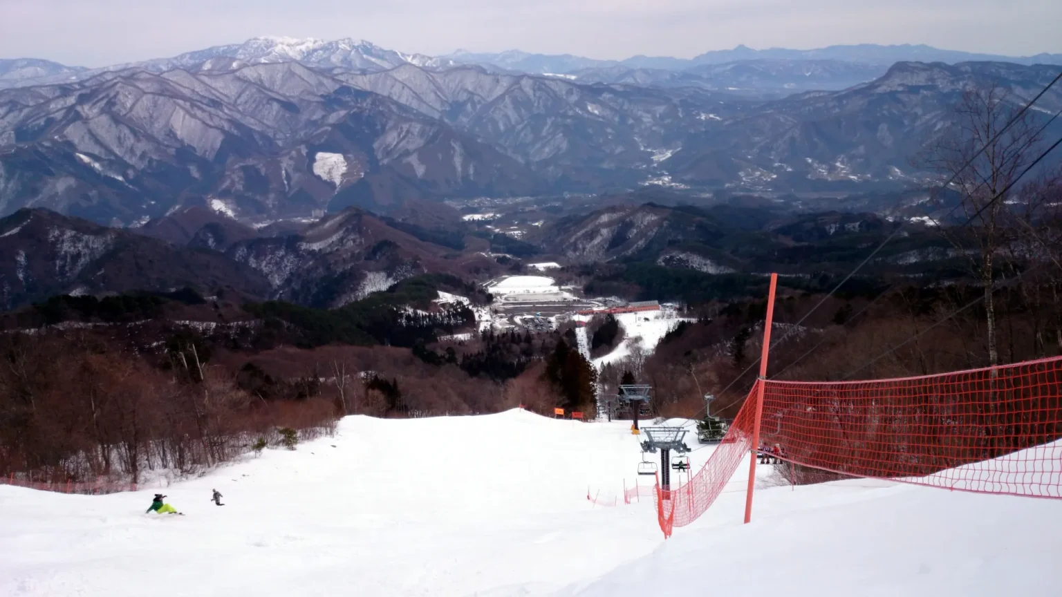 The view down a groomed piste at Norn Minakami, with an orange safety net on the right, a chairlift below, and ranges of snow-dusted mountains filling the horizon.