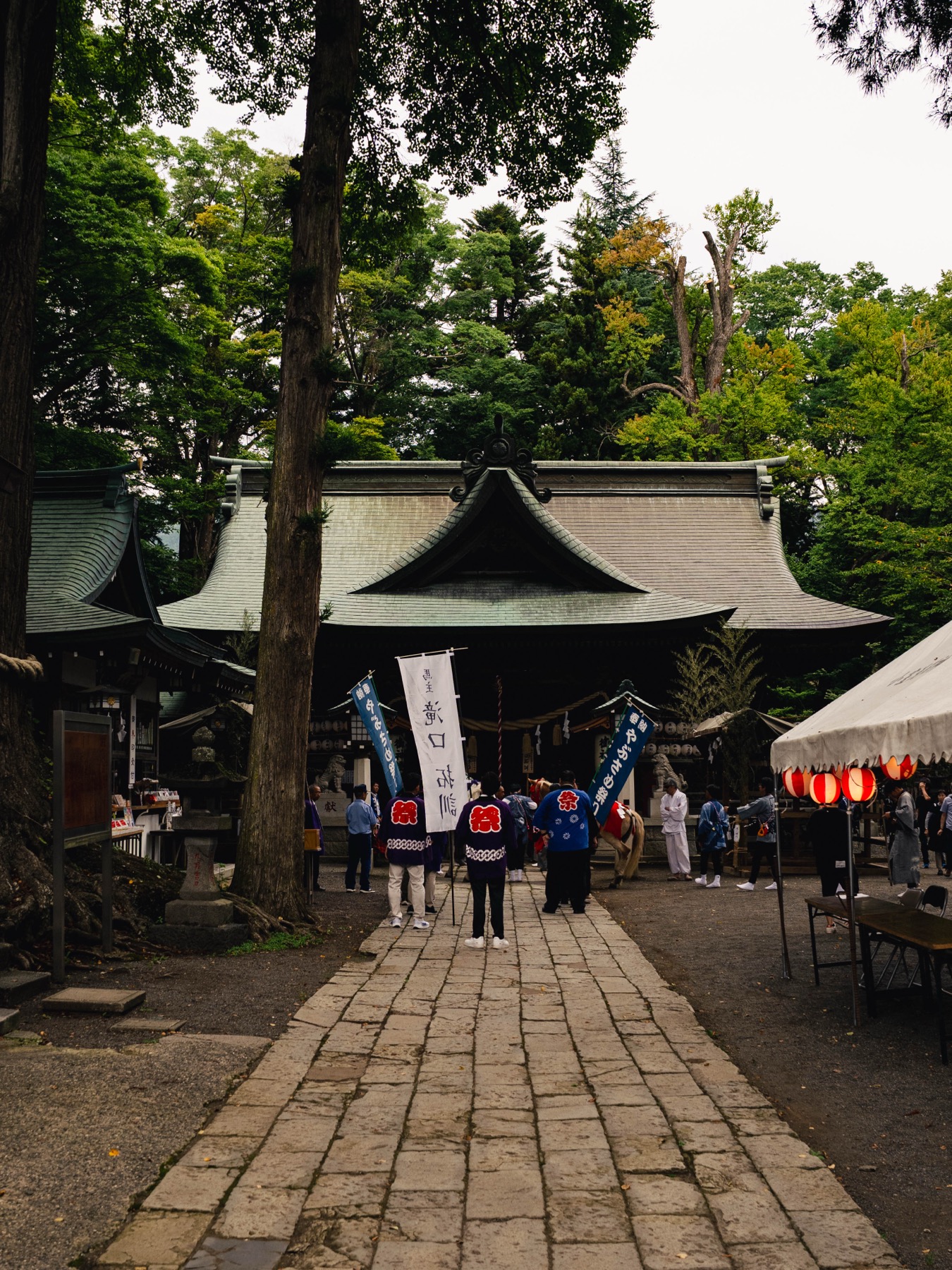 Festival morning at Omuro Sengen Shrine in Shimoyoshida: stone approach lined with food-stall tents, red lanterns and neighborhood association banners under tall cedar trees