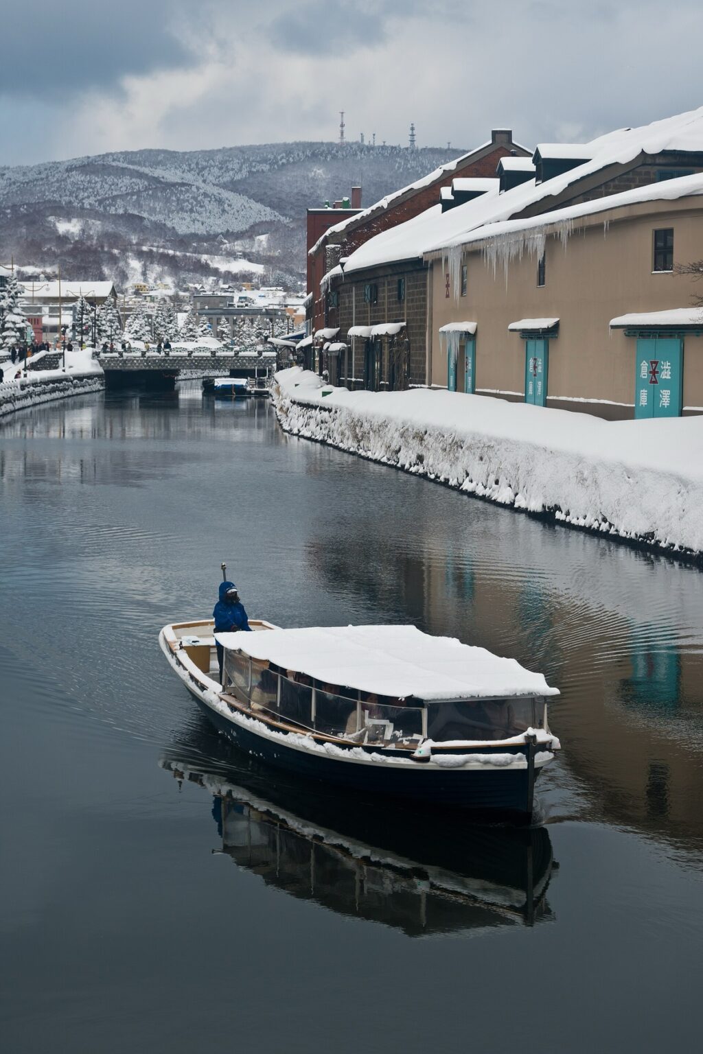 A small cruise boat with a snow-covered canopy gliding along the Otaru Canal in winter, passing the historic stone warehouses whose roofs are deep in snow, with snowy hills rising behind the town