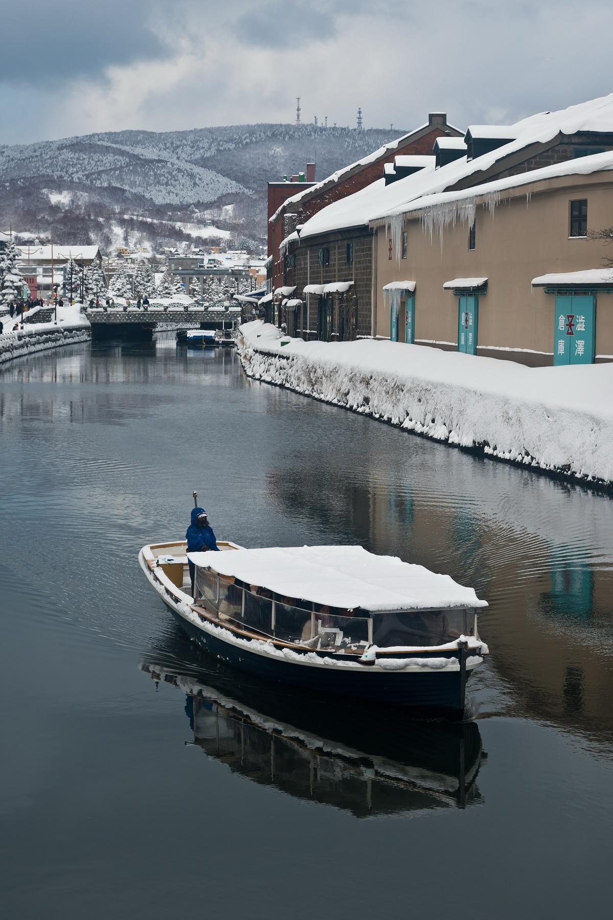 A small cruise boat with a snow-covered canopy gliding along the Otaru Canal in winter, passing the historic stone warehouses whose roofs are deep in snow, with snowy hills rising behind the town.