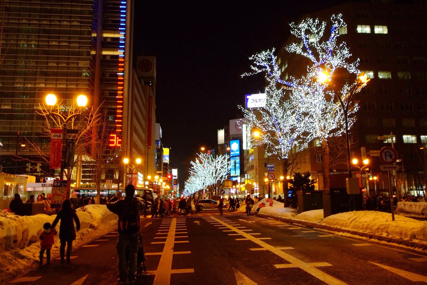 Sapporo Ekimae-dori boulevard at night during the Snow Festival period, with trees wrapped in white illuminations, snow piled along the roadside and a festival banner showing the event dates on a lamp post.