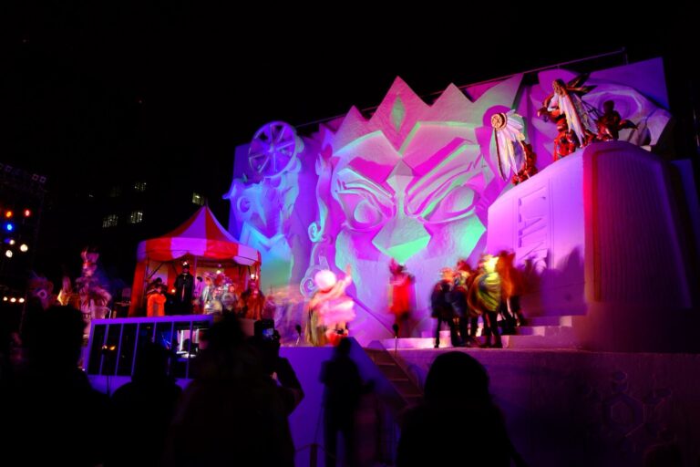 A five-storey snow sculpture at the Sapporo Snow Festival Odori site lit up at night by a colourful projection-mapping show, with costumed performers on the stage below and spectators photographing the scene.