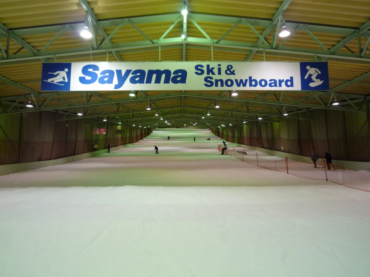 The interior of the Sayama indoor ski hall: a wide white artificial snow slope under a yellow steel roof, with skiers and snowboarders descending beneath a large Sayama Ski and Snowboard sign.