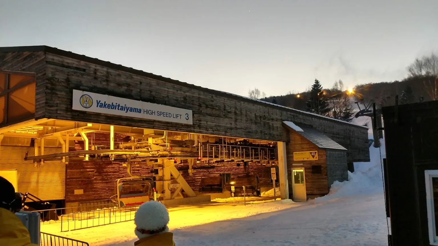 The wooden base station of the Yakebitaiyama High Speed Lift 3 at dusk, its interior lit warm yellow, with floodlights coming on above the snow behind it.