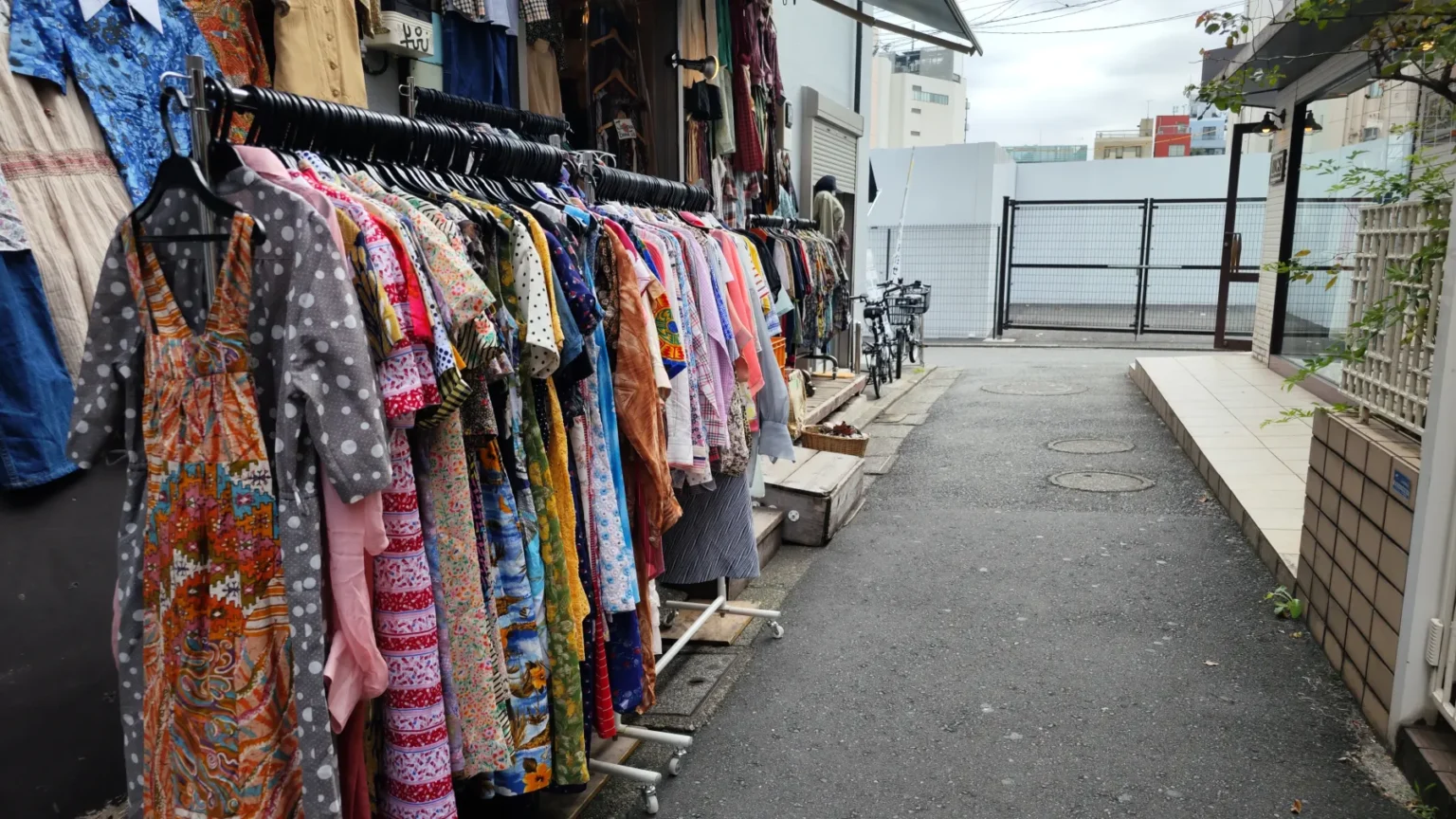 A rail of printed dresses set out along a narrow Shimokitazawa alley, with a concrete wall opposite and the passage running away from the camera.