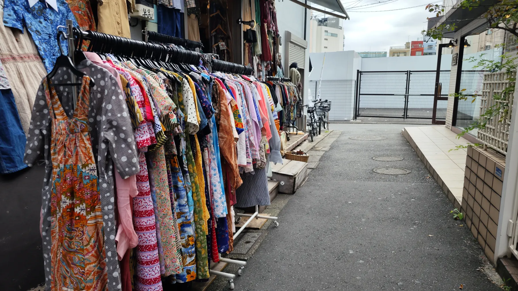 A rail of printed dresses set out along a narrow Shimokitazawa alley, with a concrete wall opposite and the passage running away from the camera.