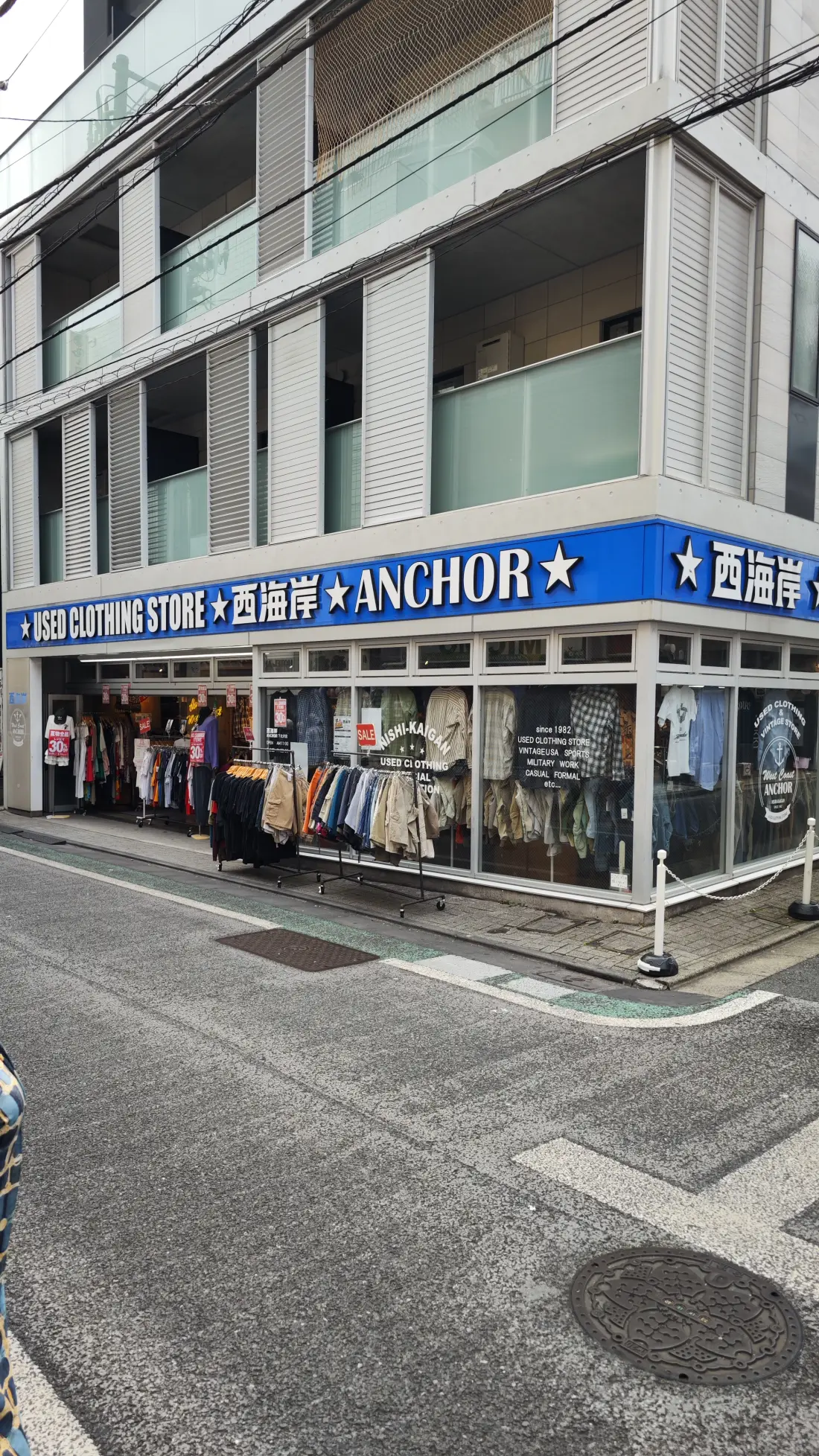 A corner building in Shimokitazawa with a long blue signboard reading Used Clothing Store Anchor, above a shopfront of stacked second-hand clothing, seen from an empty crossing.