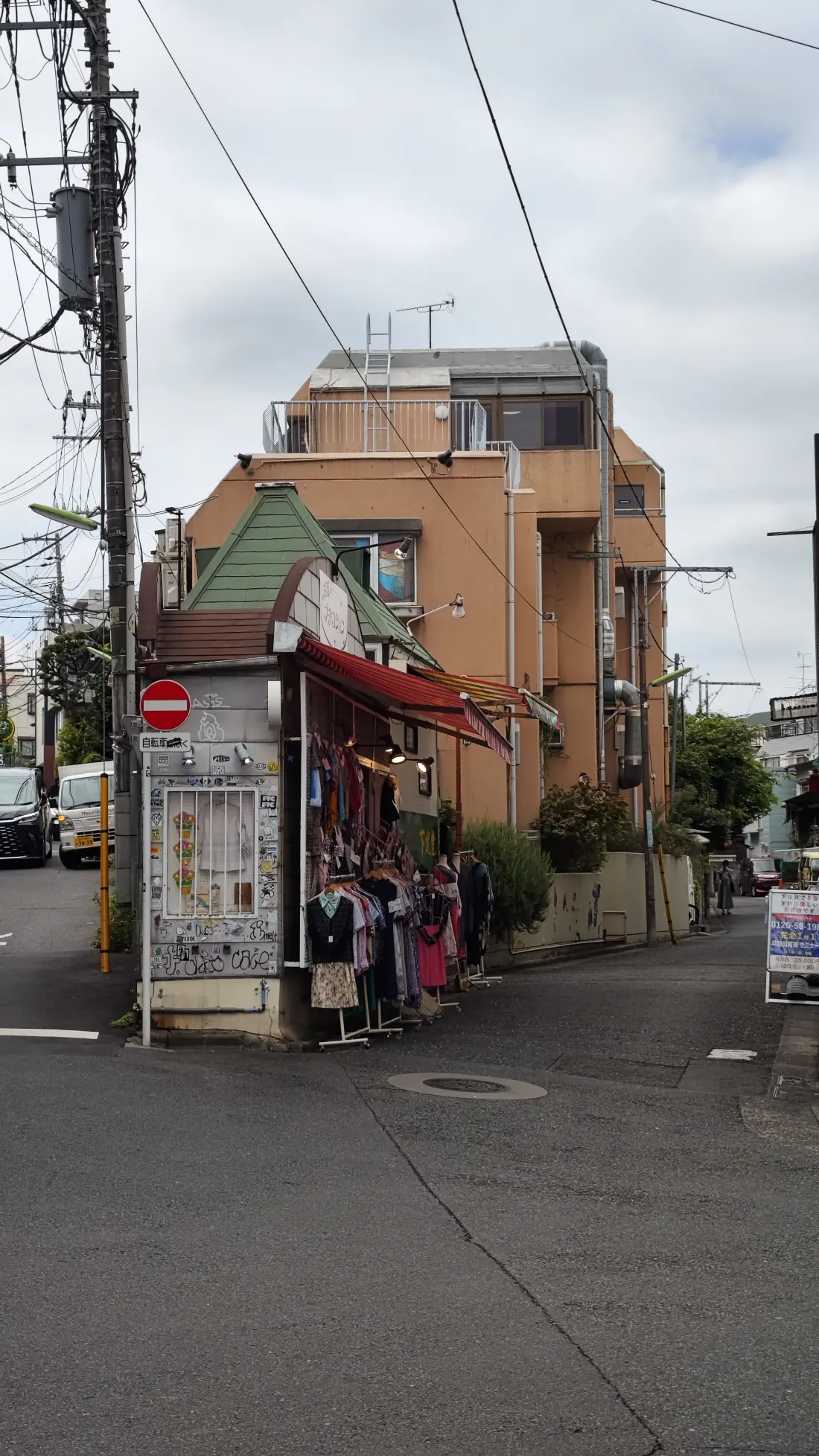 A quiet Shimokitazawa back street in the early evening, with a small shop showing a clothes rack under its awning, low two-storey buildings and overhead power lines crossing the sky.