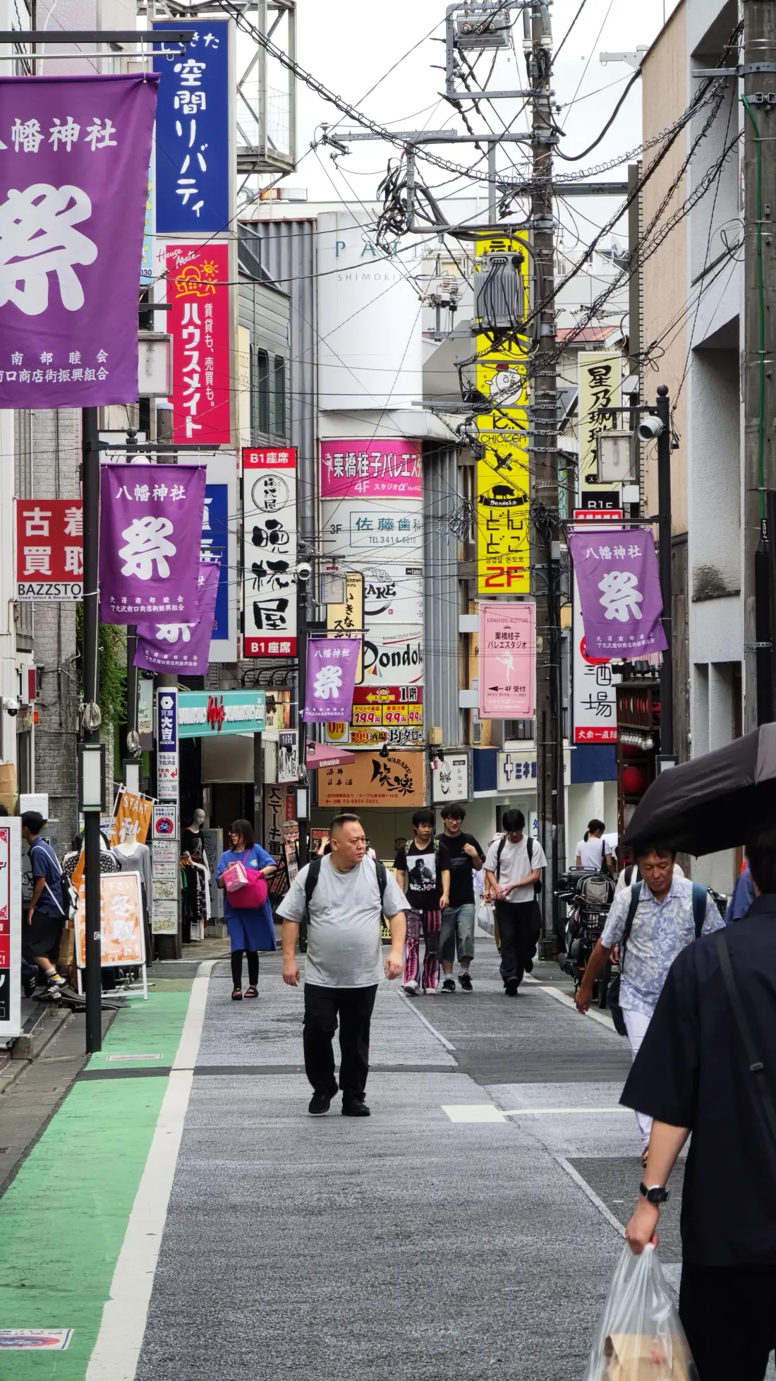 A Shimokitazawa shopping street lined on both sides with purple festival banners and vertical shop signs, with shoppers walking along the green-edged pavement.