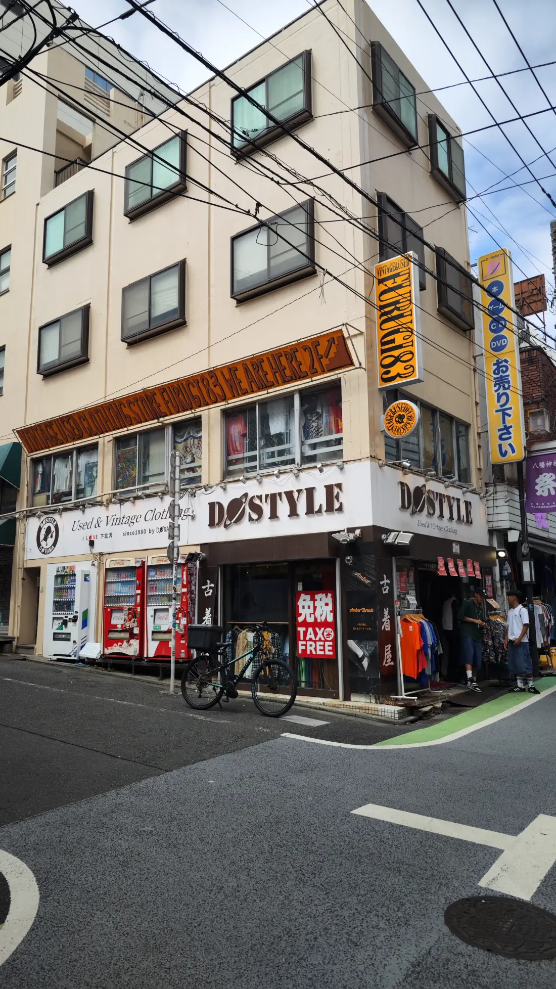 A wider view of a Shimokitazawa street corner with the DOSTYLE vintage-clothing shop on the ground floor, a parked bicycle and apartment floors stacked above it.