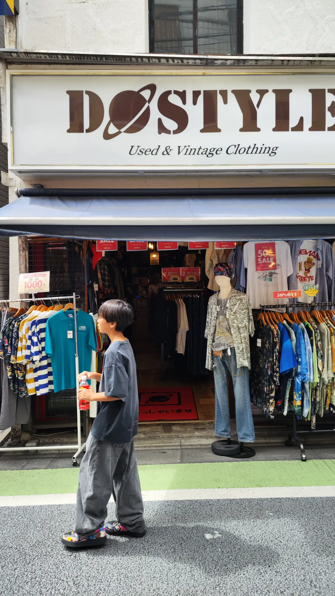 The frontage of a Shimokitazawa vintage shop with a large sign reading DOSTYLE Used and Vintage Clothing, red sale notices in the window and a shopper looking in.