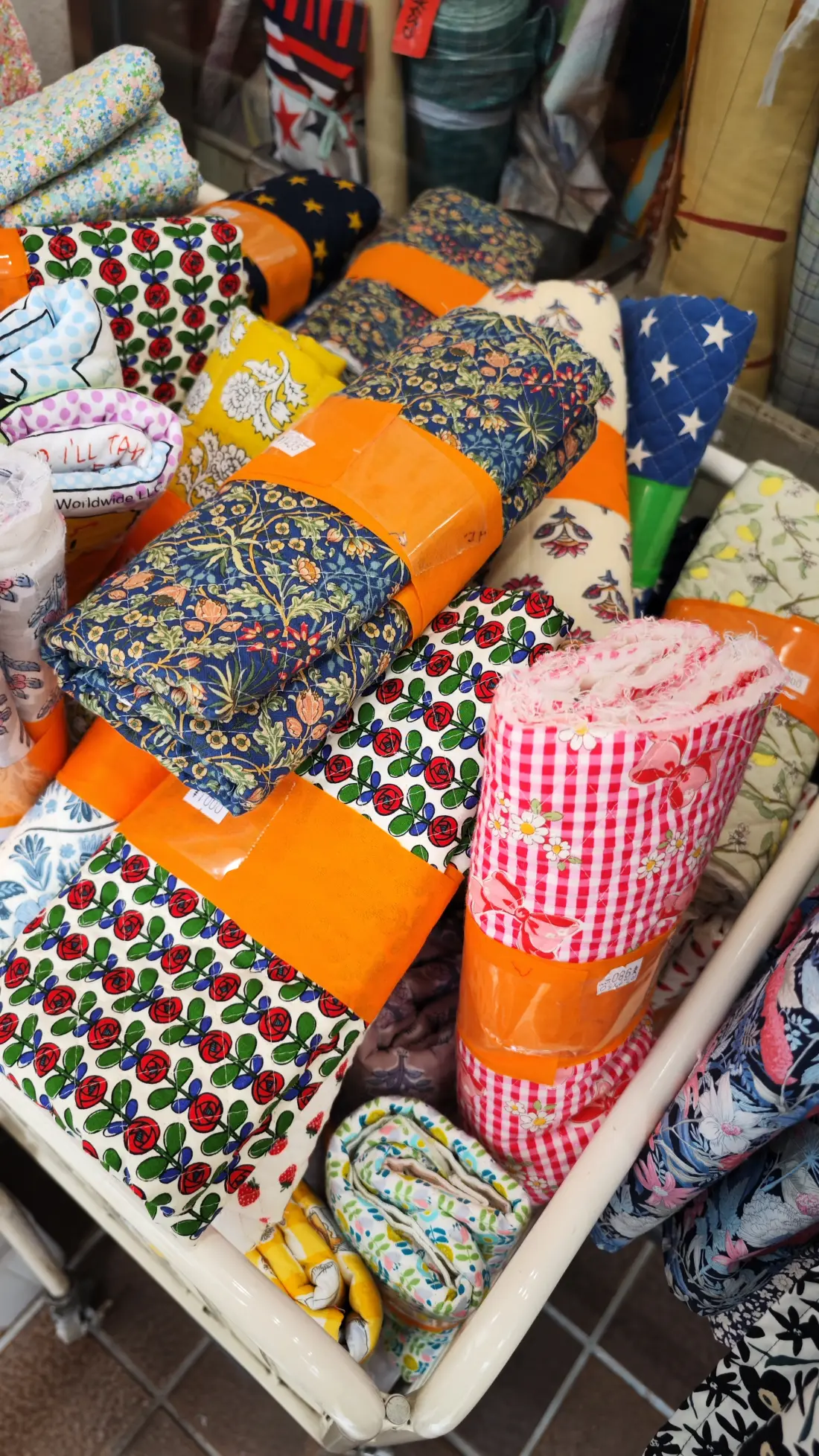 Rolls of printed cotton fabric wrapped in orange tape and stacked outside a Shimokitazawa fabric shop, with handwritten price tags attached to them.