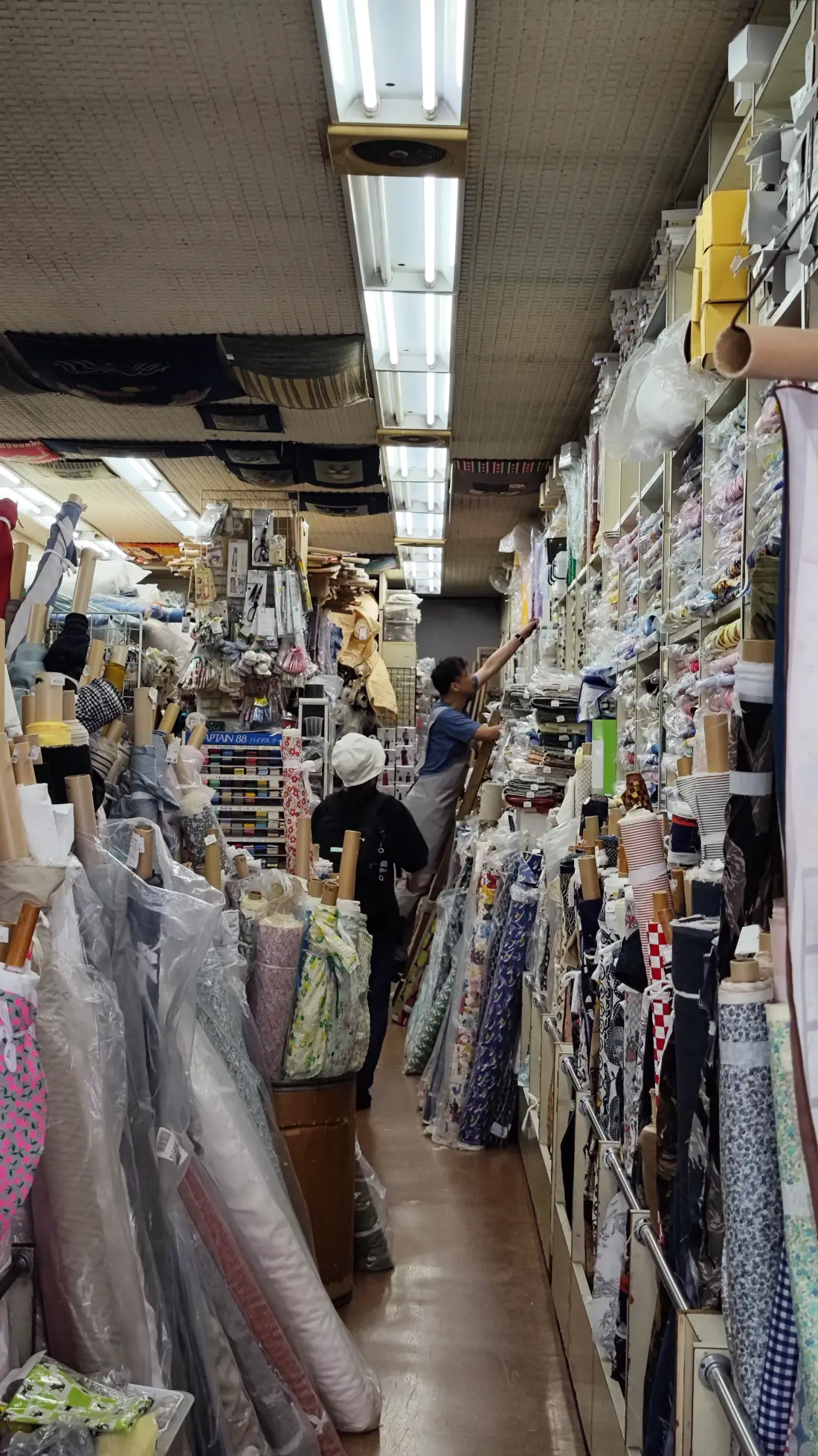 A narrow aisle inside a Shimokitazawa fabric shop, with bolts of cloth in plastic sleeves stacked from floor to ceiling under fluorescent lights.
