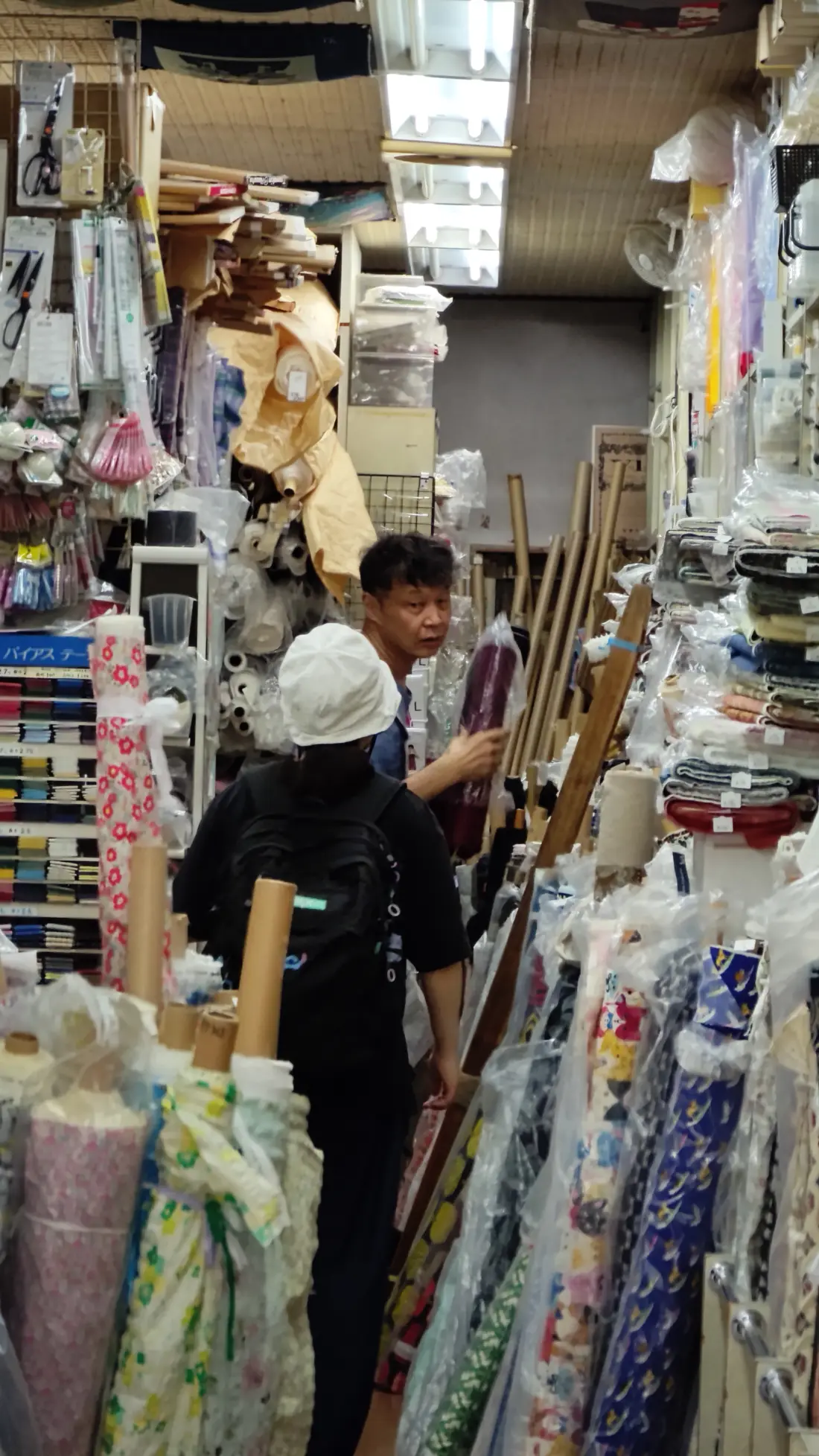 A person looking through densely stacked bolts of fabric inside a crowded Shimokitazawa fabric shop, with rolls filling every shelf around them.