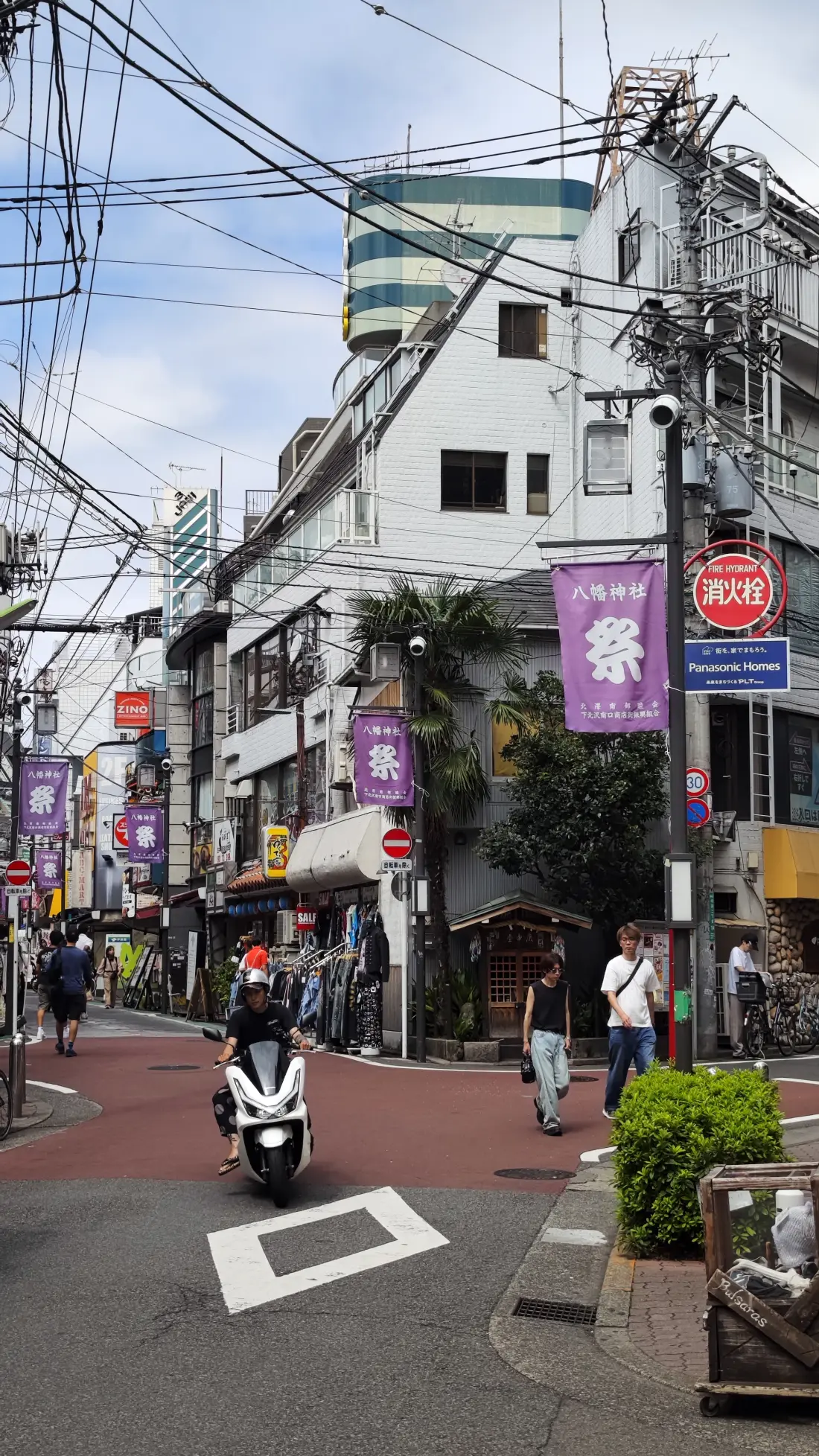 A narrow Shimokitazawa street paved in reddish asphalt, with purple Hachiman Shrine festival banners hanging from poles, a parked scooter and dense overhead cables above.