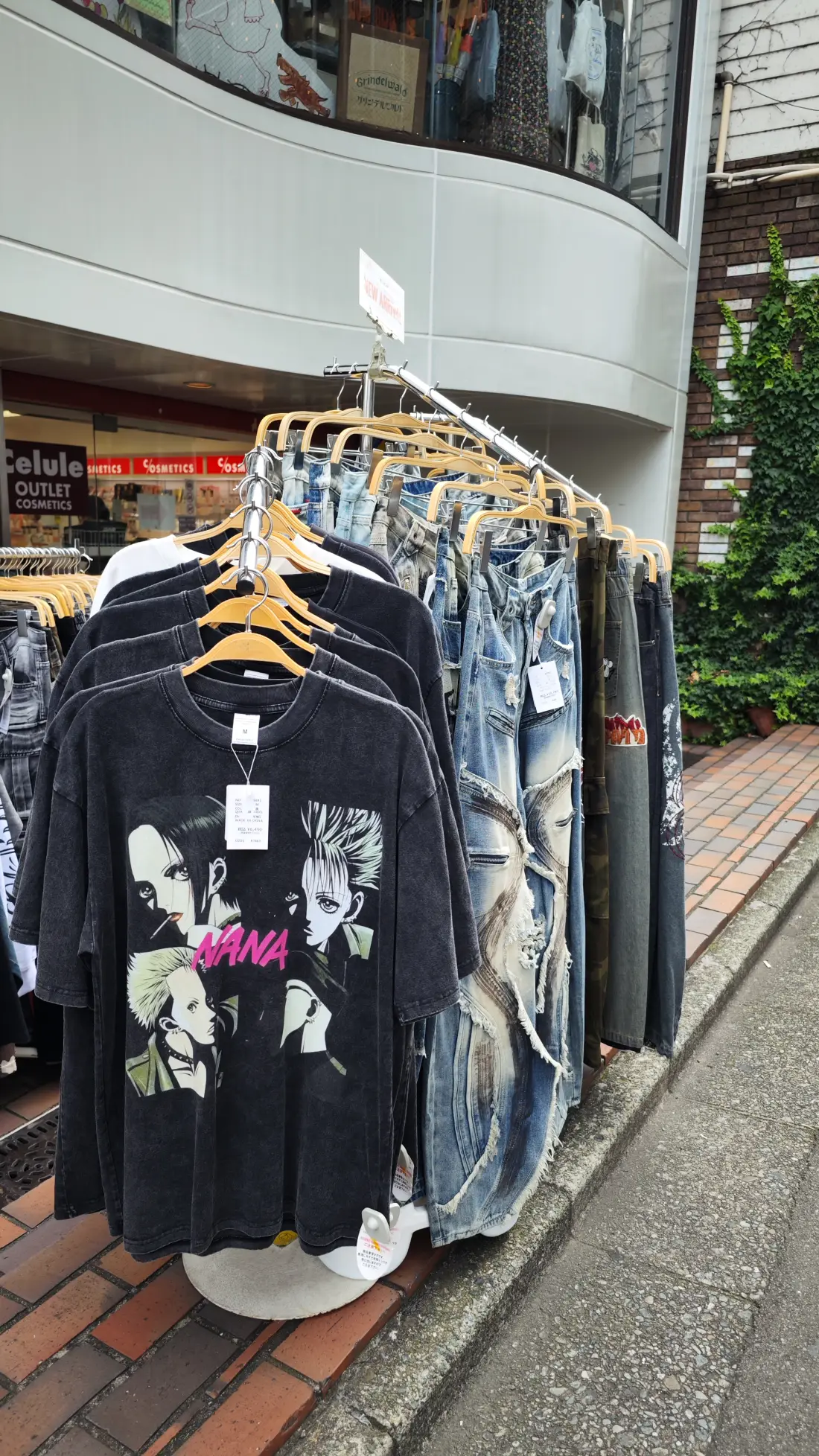 A black printed T-shirt on a wooden hanger displayed outdoors in Shimokitazawa, with denim garments hanging behind it above brick paving.