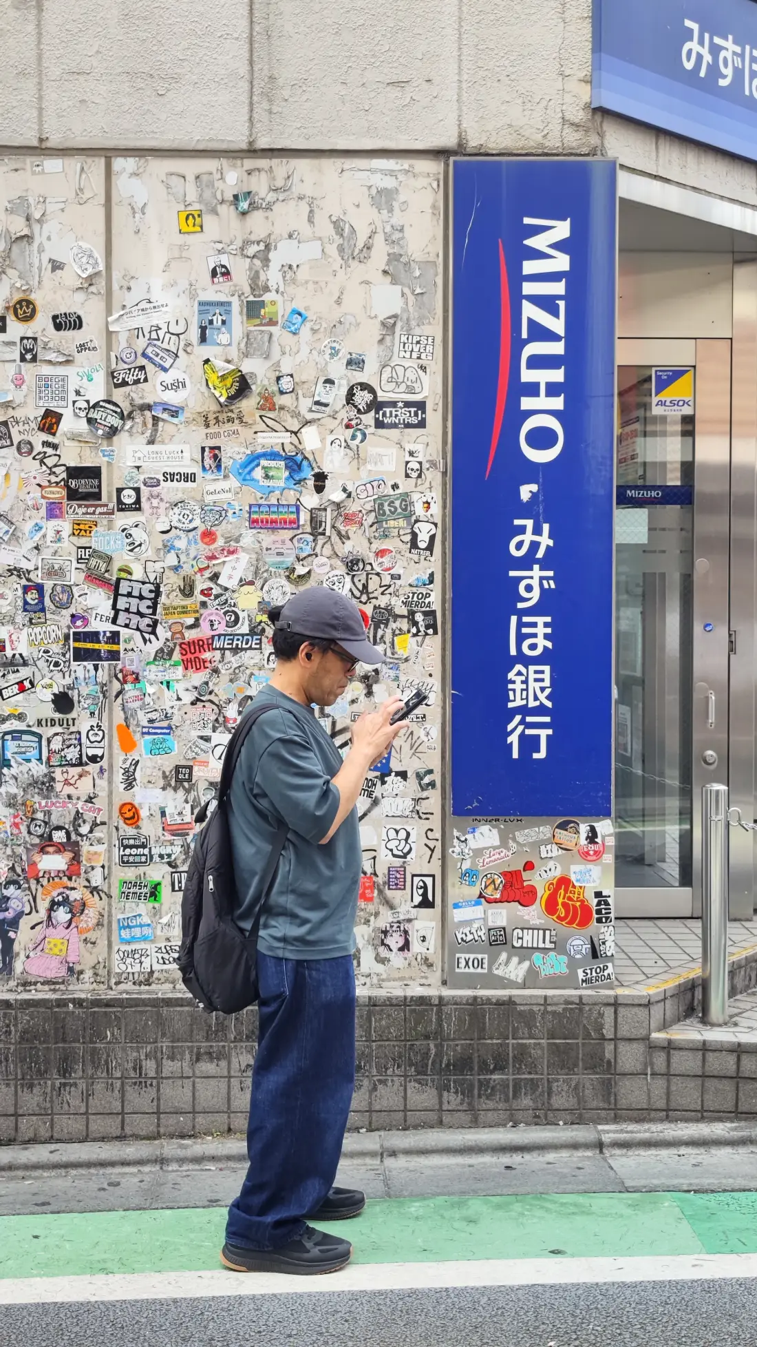 A person standing beside a Shimokitazawa bank wall that has been completely covered in stickers, next to a blue Mizuho Bank sign at street level.