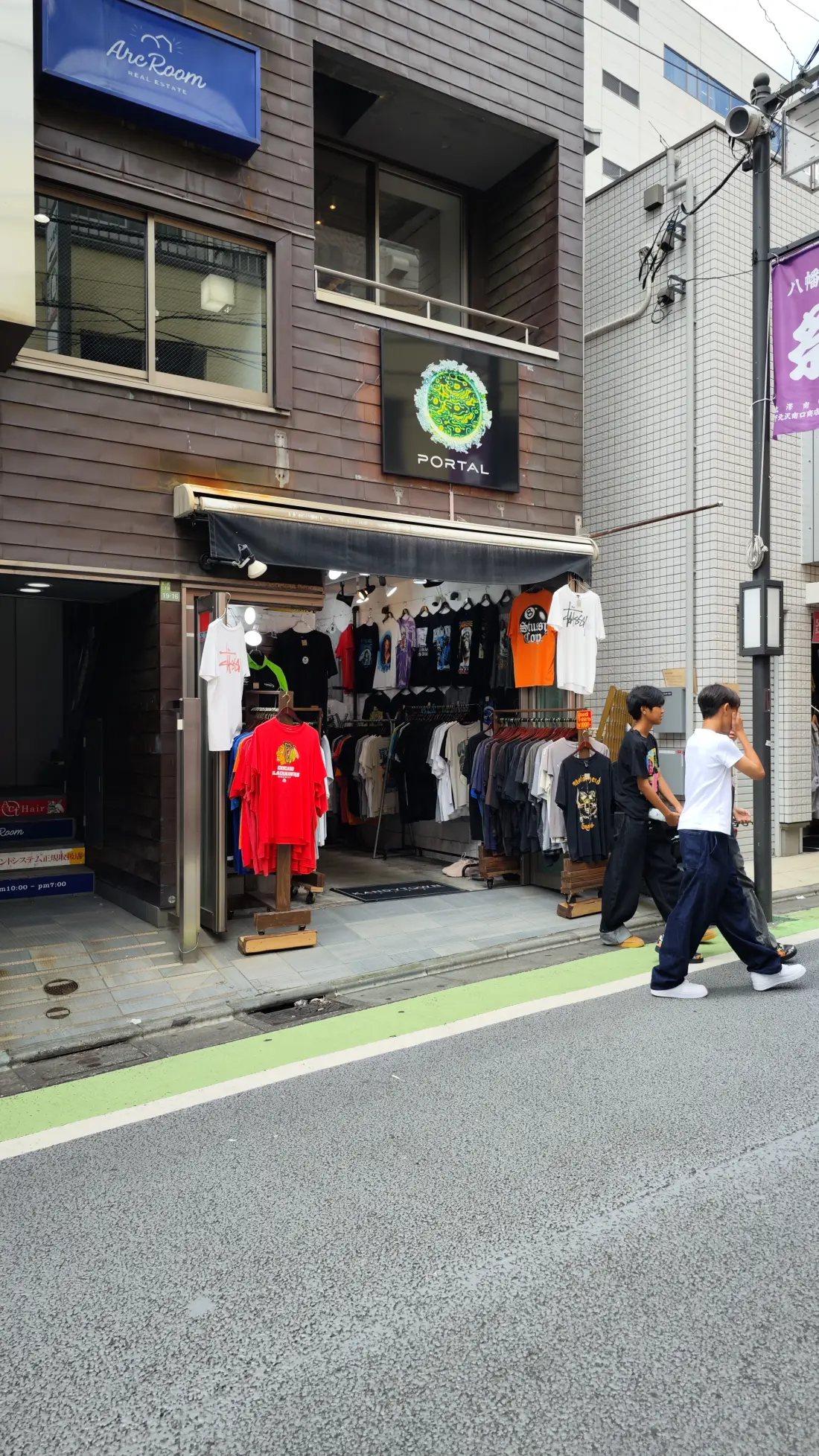 A narrow Shimokitazawa lane with a clothing shop open to the street, garments hung along the frontage and pedestrians walking along the green-edged roadway.