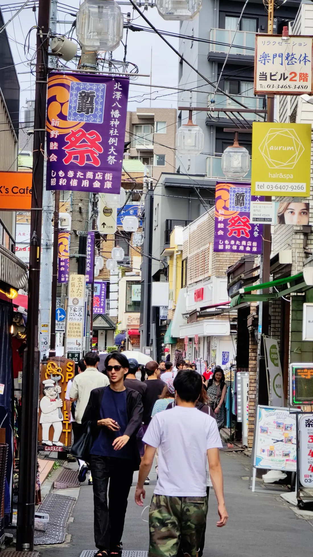 A dense Shimokitazawa side street packed with illuminated vertical signs for bars and restaurants, a purple festival banner overhead and people walking through it.