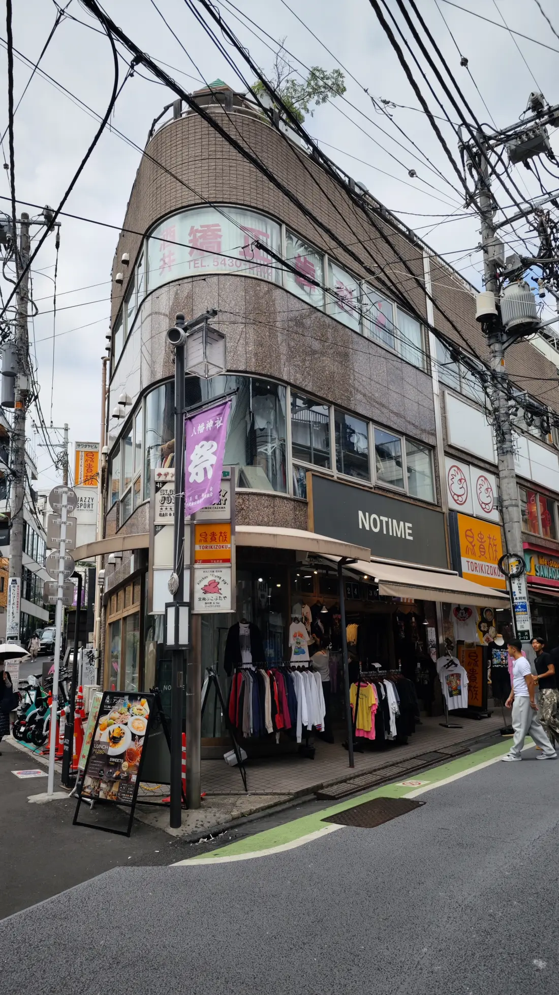 A curved corner building in Shimokitazawa housing a vintage-clothing shop, with garment rails set out on the pavement and overhead cables running across the frame.