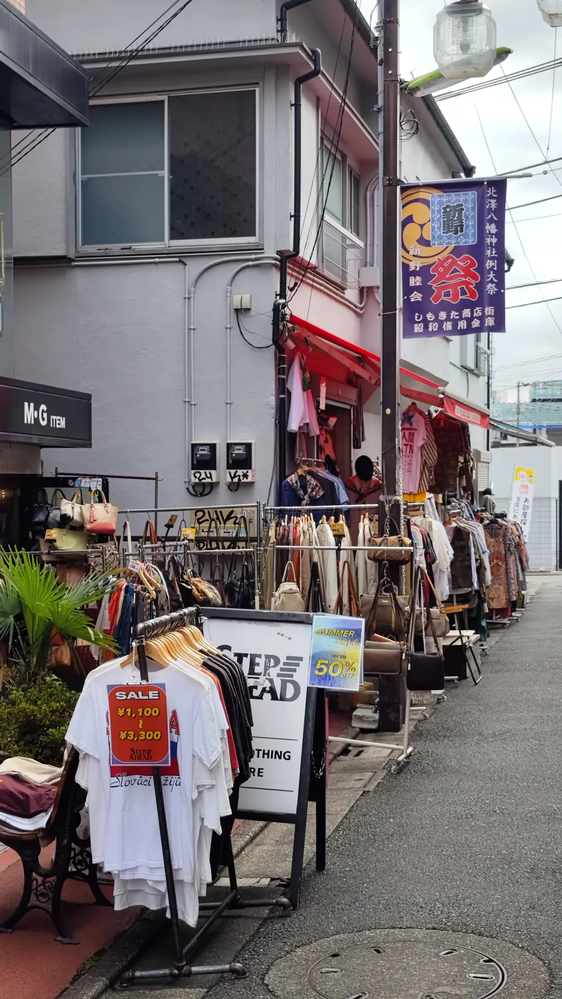 Clothing rails set out on a Shimokitazawa street corner with a red sale notice, potted plants beside the shopfront and a red and white awning above them.