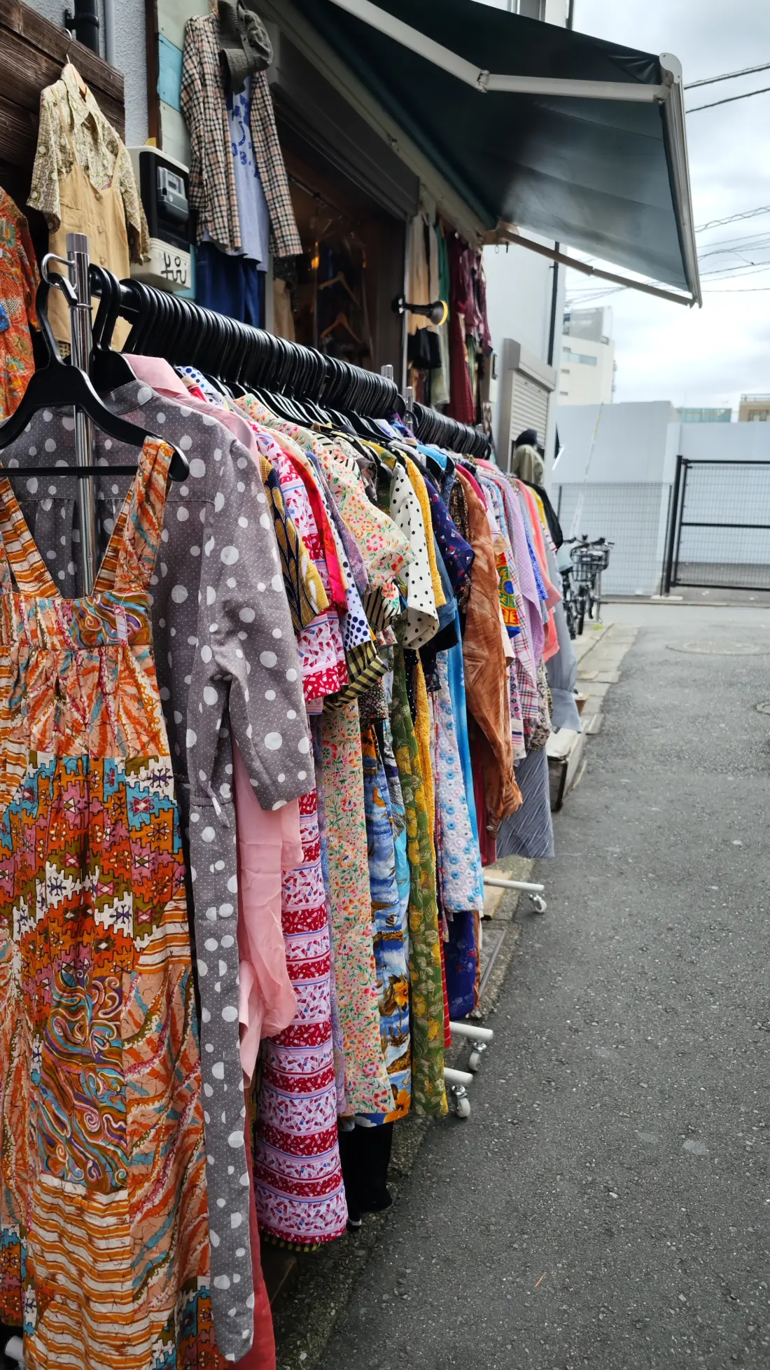 A closely packed rail of brightly printed dresses hanging outside a Shimokitazawa shop under a dark awning.