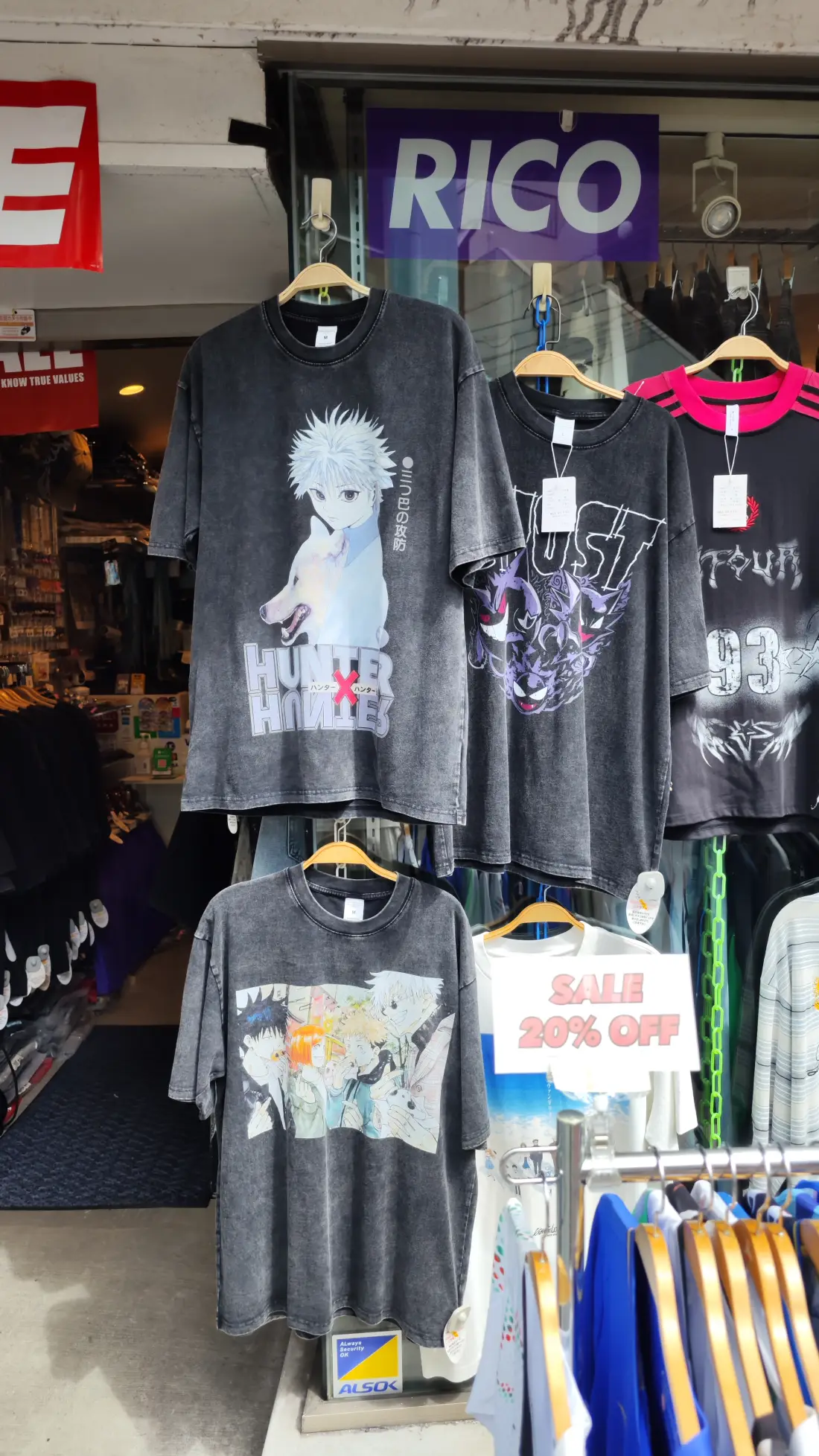 Printed graphic T-shirts hanging on a rail outside a Shimokitazawa vintage shop under a sign reading RICO, with a discount notice and folded umbrellas standing beside them.