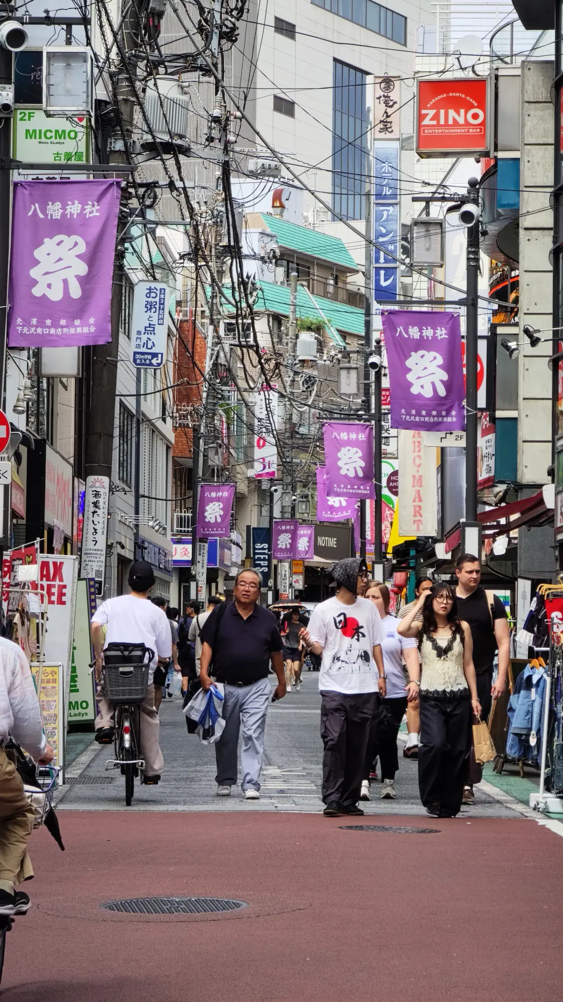 Shoppers walking down a busy Shimokitazawa shopping street lined with purple banners reading Hachiman Shrine Festival, with stacked shop signs and overhead wires above them.