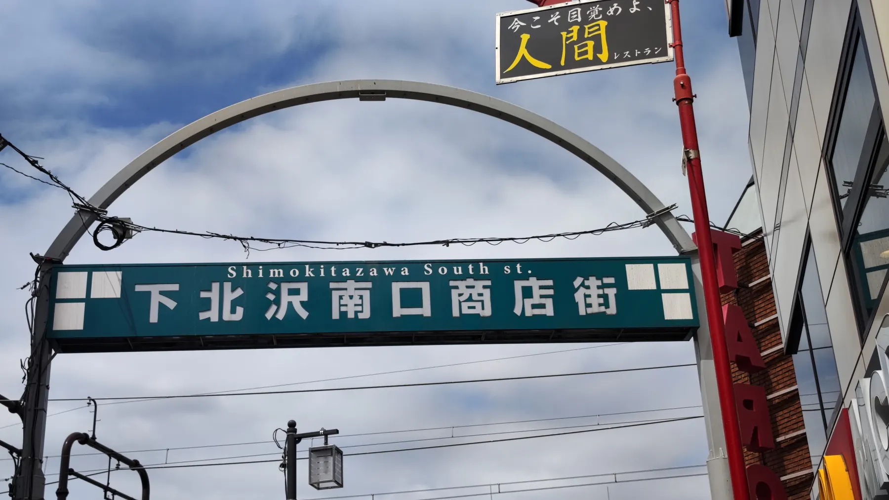 The steel entrance arch of the Shimokitazawa South Exit shopping street, with a green nameplate carrying the Japanese name and the words Shimokitazawa South st against a cloudy sky.