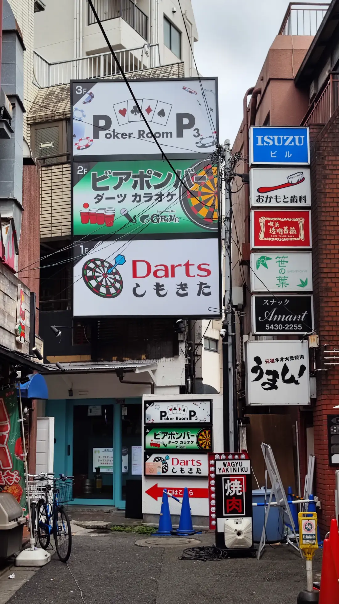 A vertical stack of illuminated shop signs on a Shimokitazawa building advertising a poker room, darts, karaoke and bars, with bicycles parked at street level below.