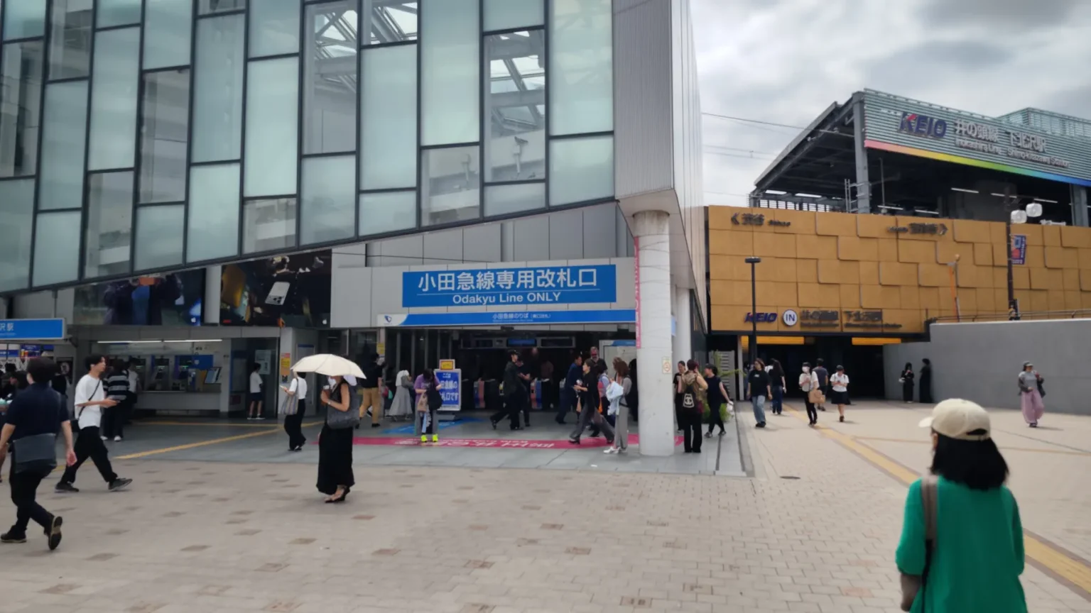 The Shimokitazawa Station forecourt with the blue Odakyu Line Only ticket gate on the left and the Keio Inokashira Line entrance on the right, and commuters walking between them.