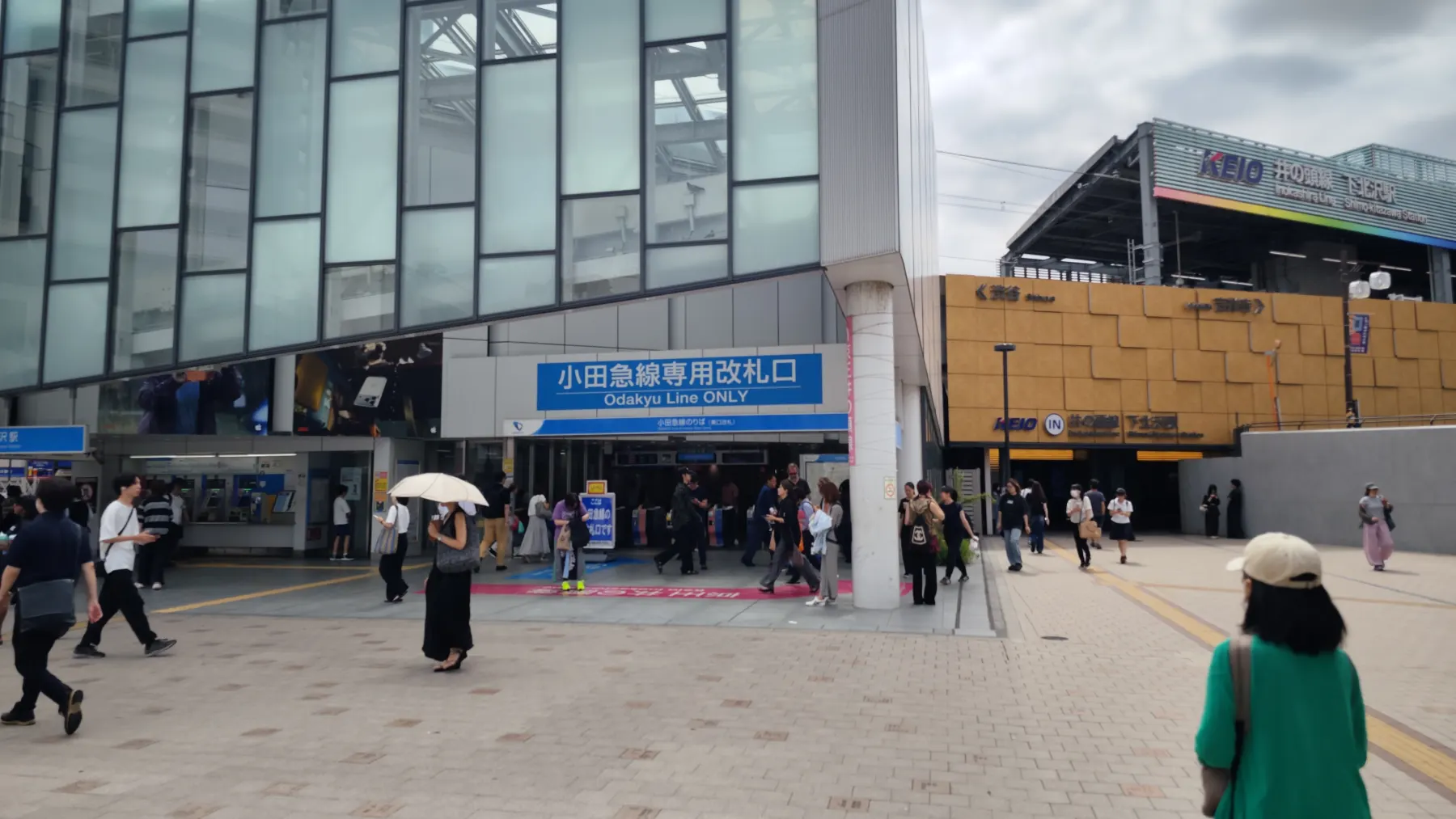 The Shimokitazawa Station forecourt with the blue Odakyu Line Only ticket gate on the left and the Keio Inokashira Line entrance on the right, and commuters walking between them.