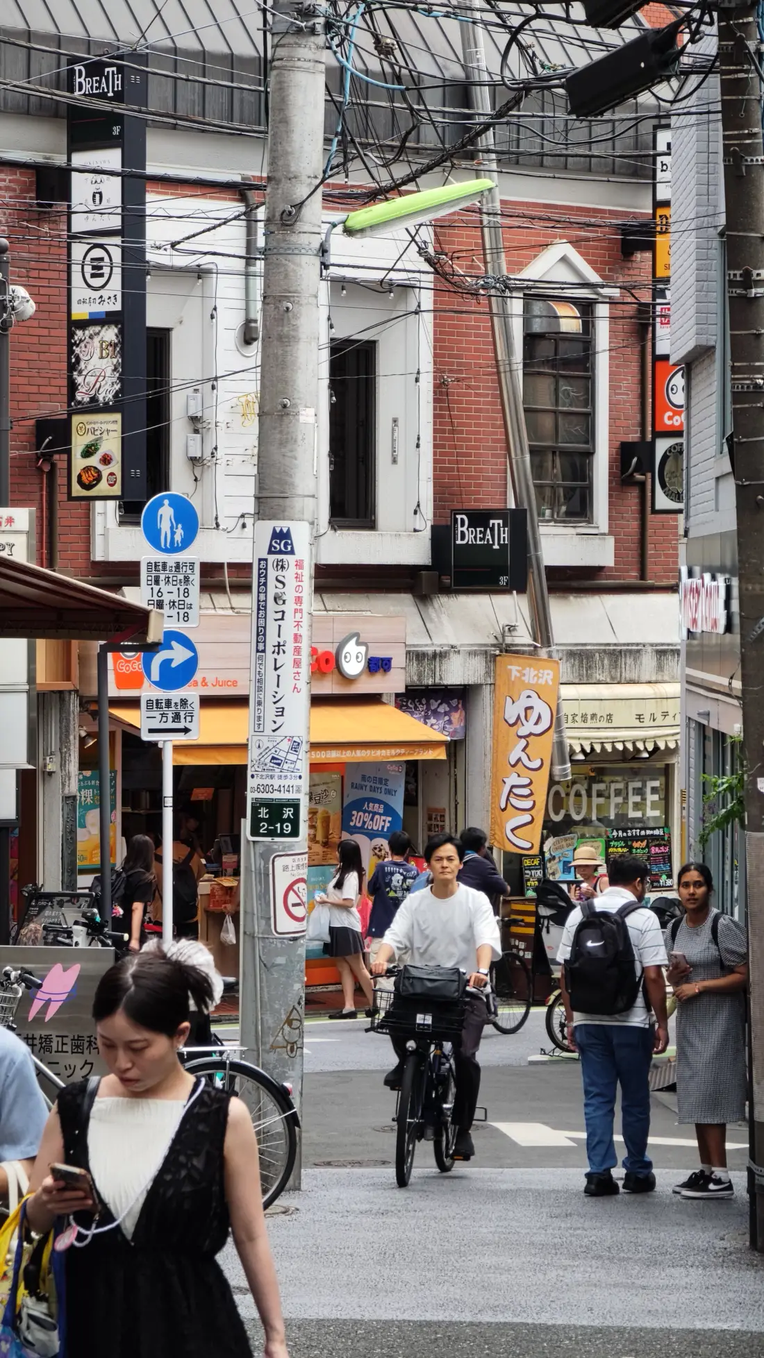 A Shimokitazawa street corner in the early evening, with a utility pole, direction signs, parked bicycles and pedestrians passing cafes and small shops.