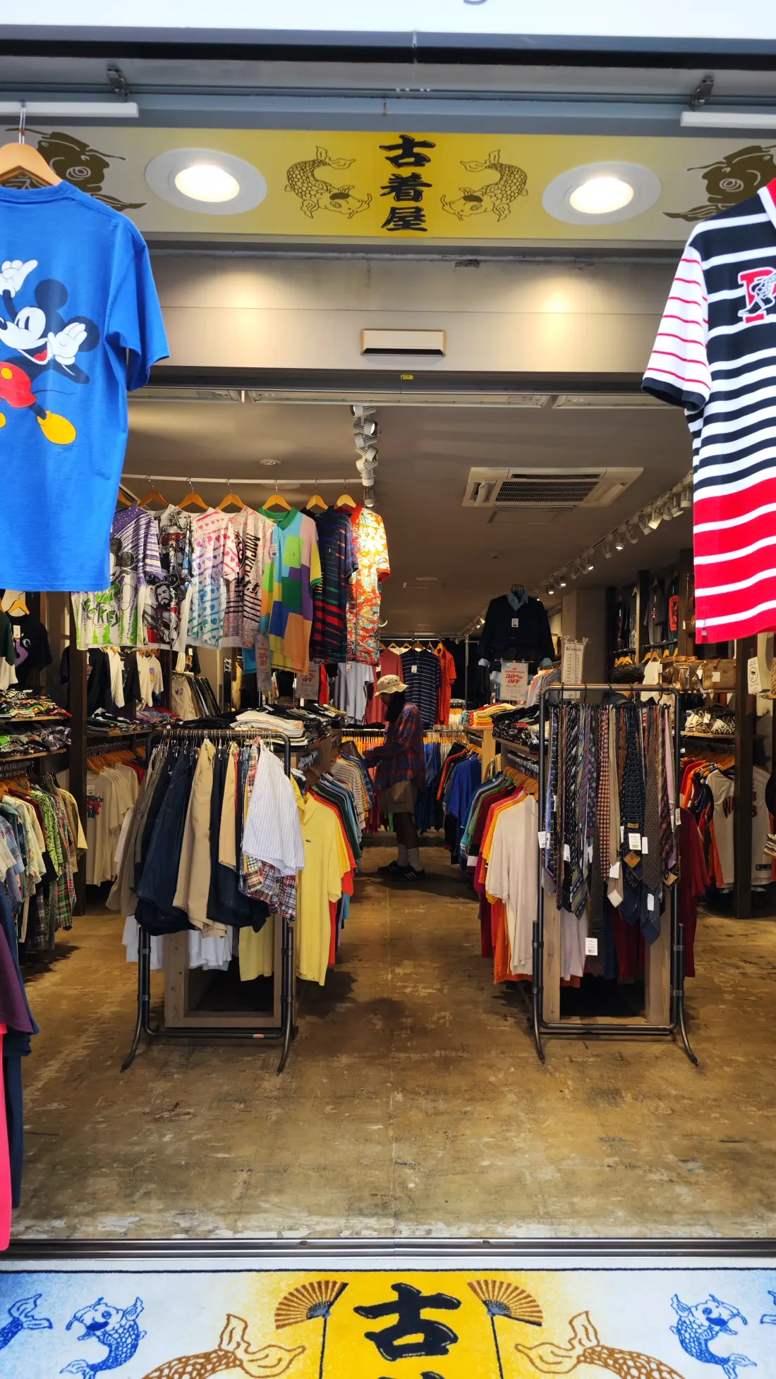 The interior of a Shimokitazawa used-clothing shop, with densely packed rails of second-hand garments running to the back and printed T-shirts displayed high on the walls.
