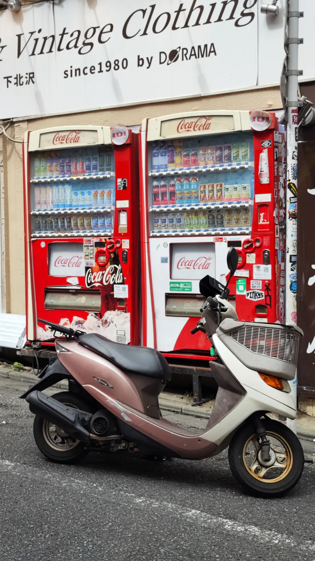 A Shimokitazawa shop sign reading Vintage Clothing since 1980 mounted above two red Coca-Cola vending machines, with a scooter parked on the pavement below it.